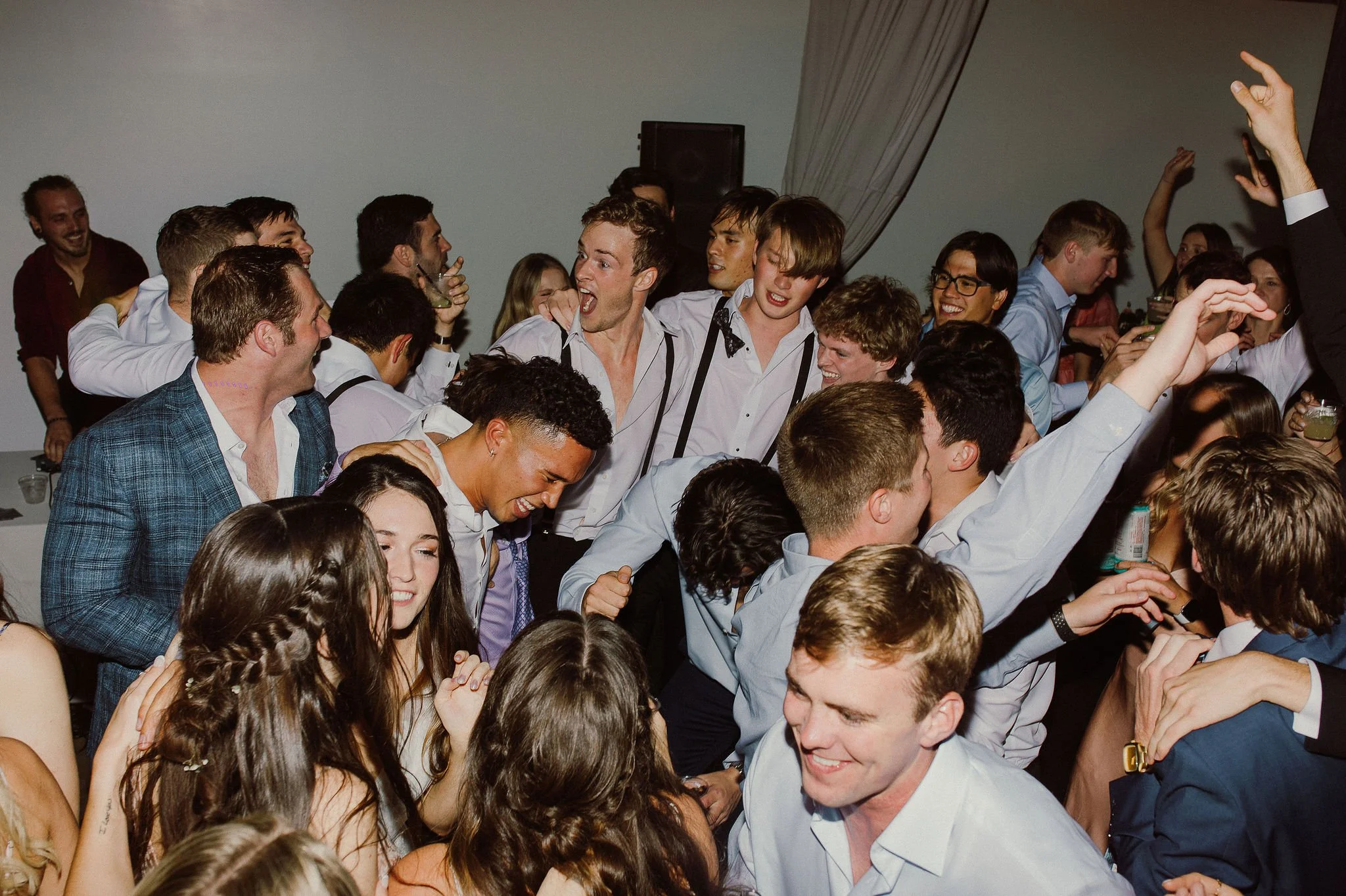 A large group of young adults at a party or celebration, dancing and enjoying themselves in a crowded indoor space.