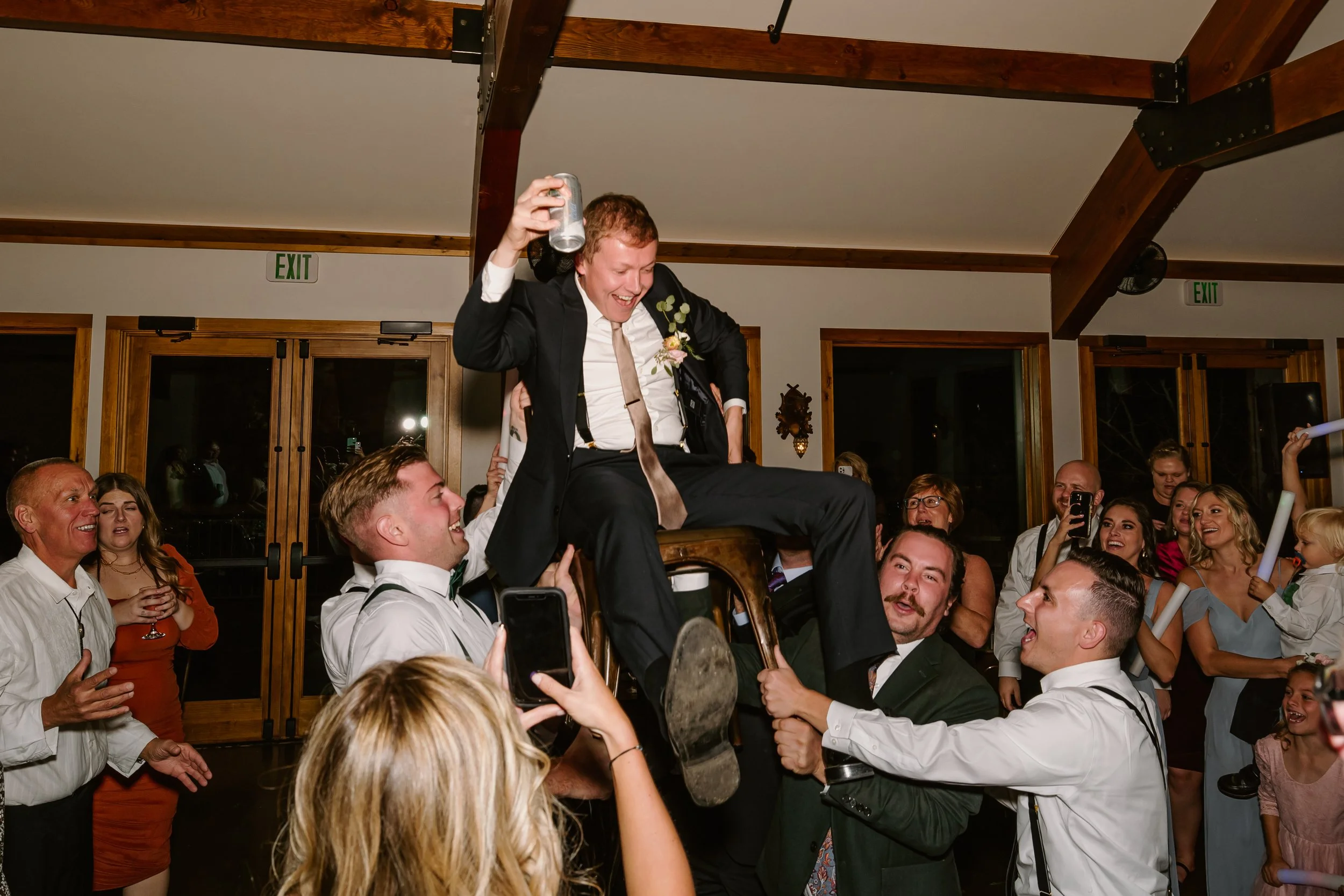 A lively celebration scene with a man in a tuxedo being lifted onto a chair by others, surrounded by smiling guests in a decorated indoor venue.
