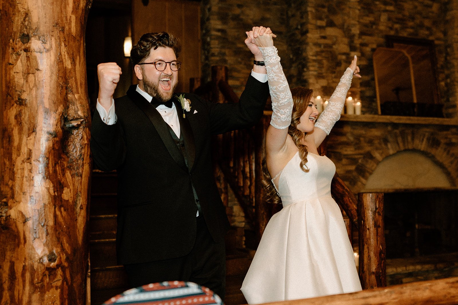 A newlywed couple celebrating at their wedding reception, with the groom in a tuxedo and the bride in a white wedding gown, both raising their arms in excitement and joy.