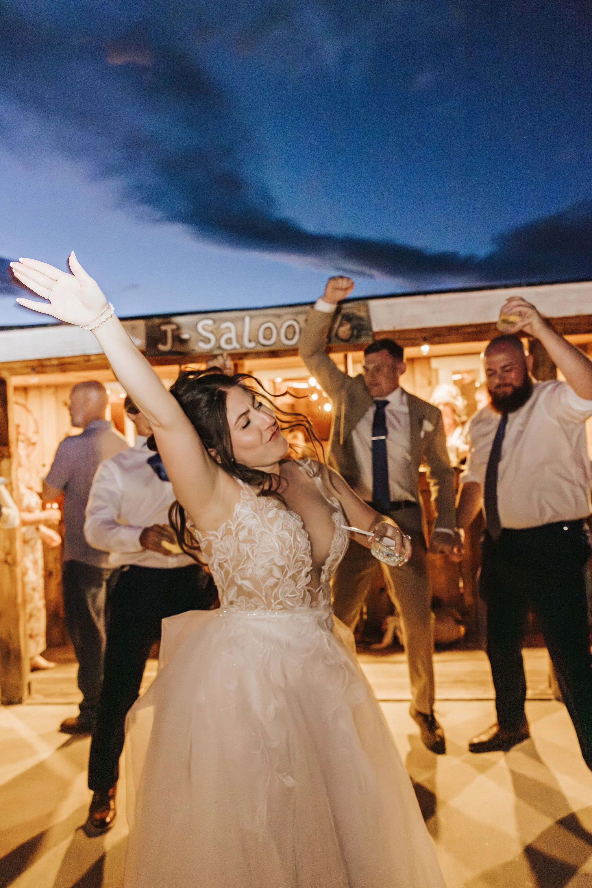A woman in a wedding dress dancing at a celebration, surrounded by people in formal attire, indoors with warm lighting and a wooden sign that says "J. Saloo".