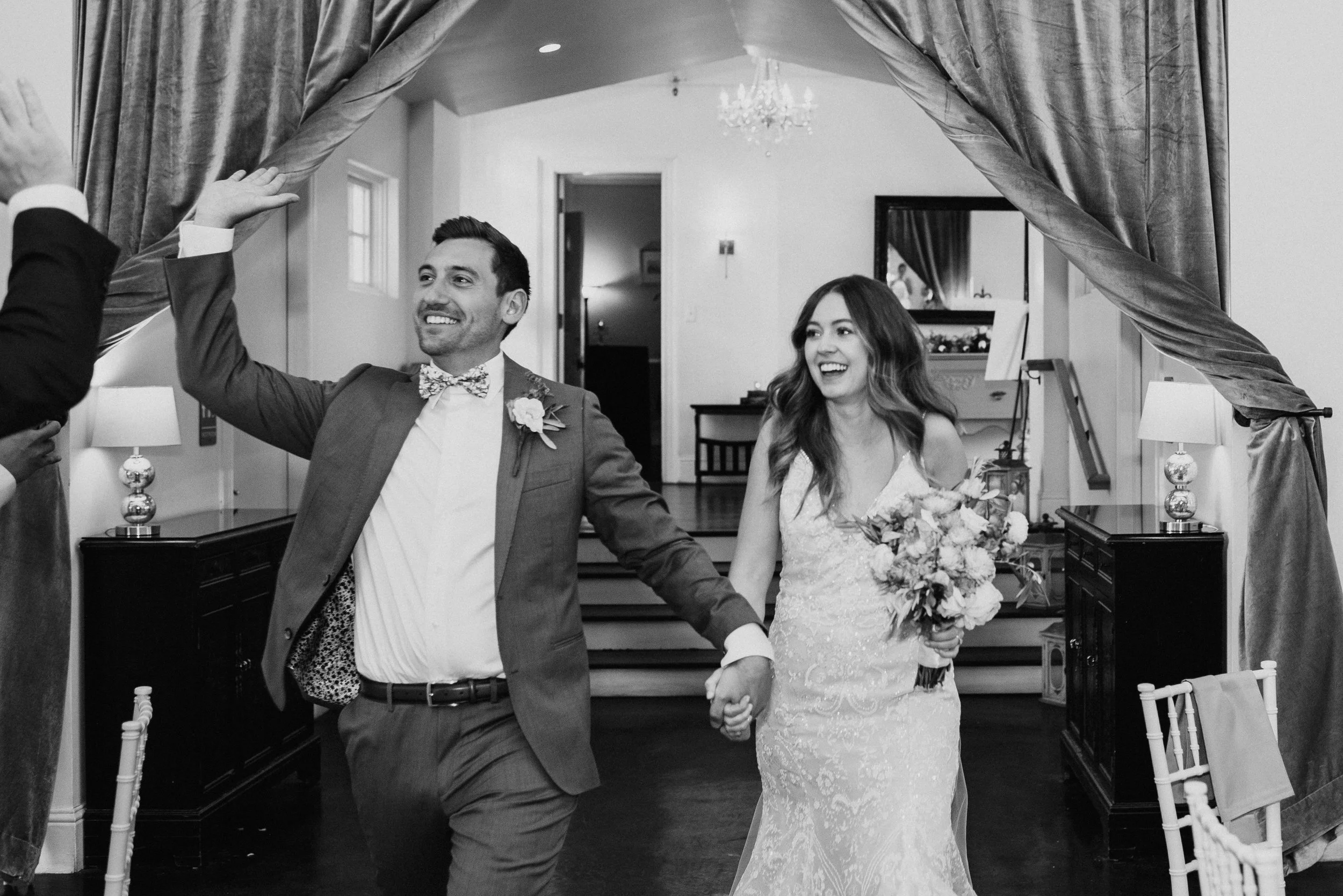 A newlywed couple holding hands and smiling while walking through a decorated doorway during their wedding reception, with a man officiating or greeting them. The groom is wearing a suit with a bowtie, and the bride is wearing a wedding dress and holding a bouquet of flowers.
