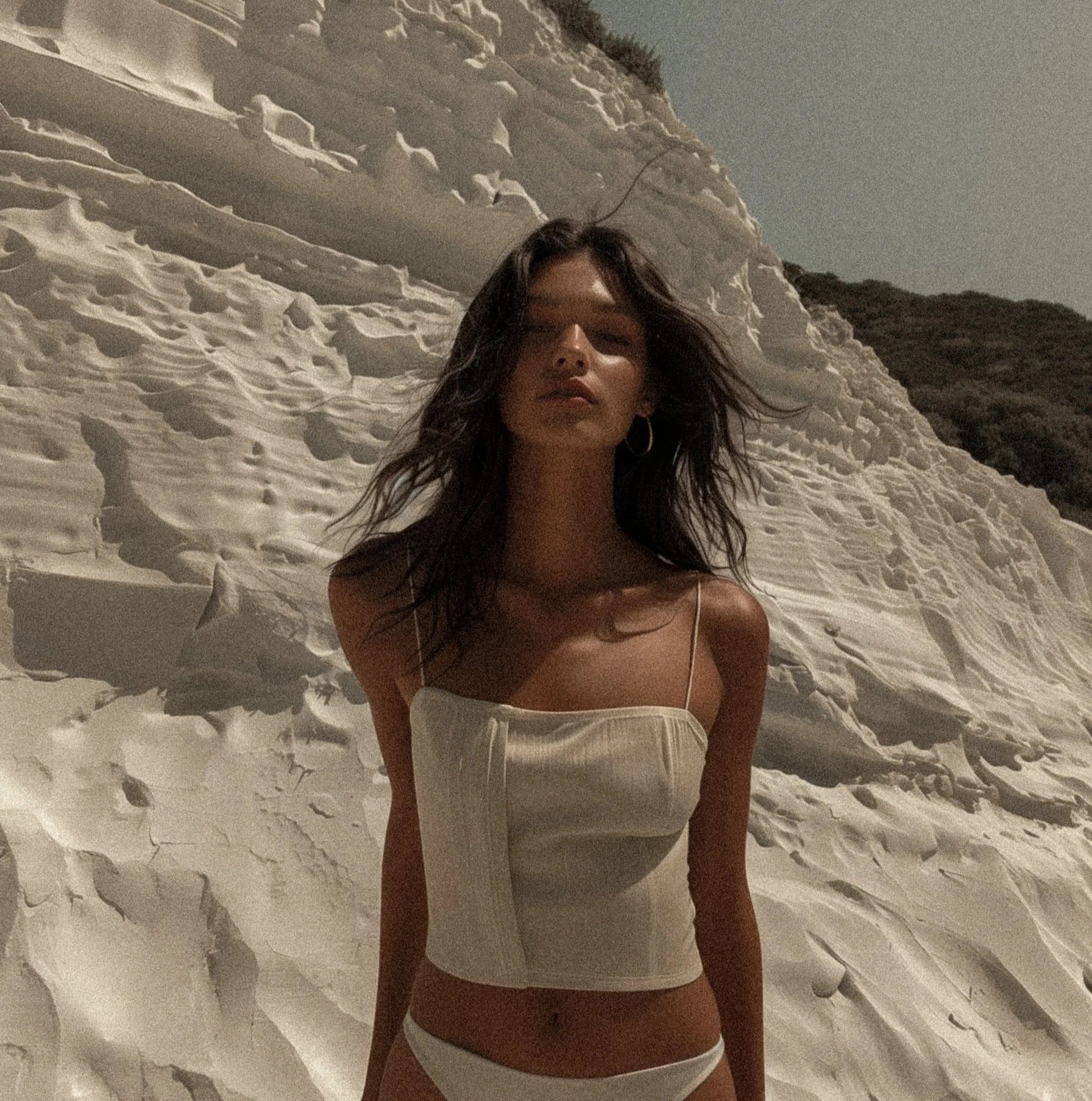 Woman with long hair and hoop earrings standing in white sand with a large sand dune behind her at night.