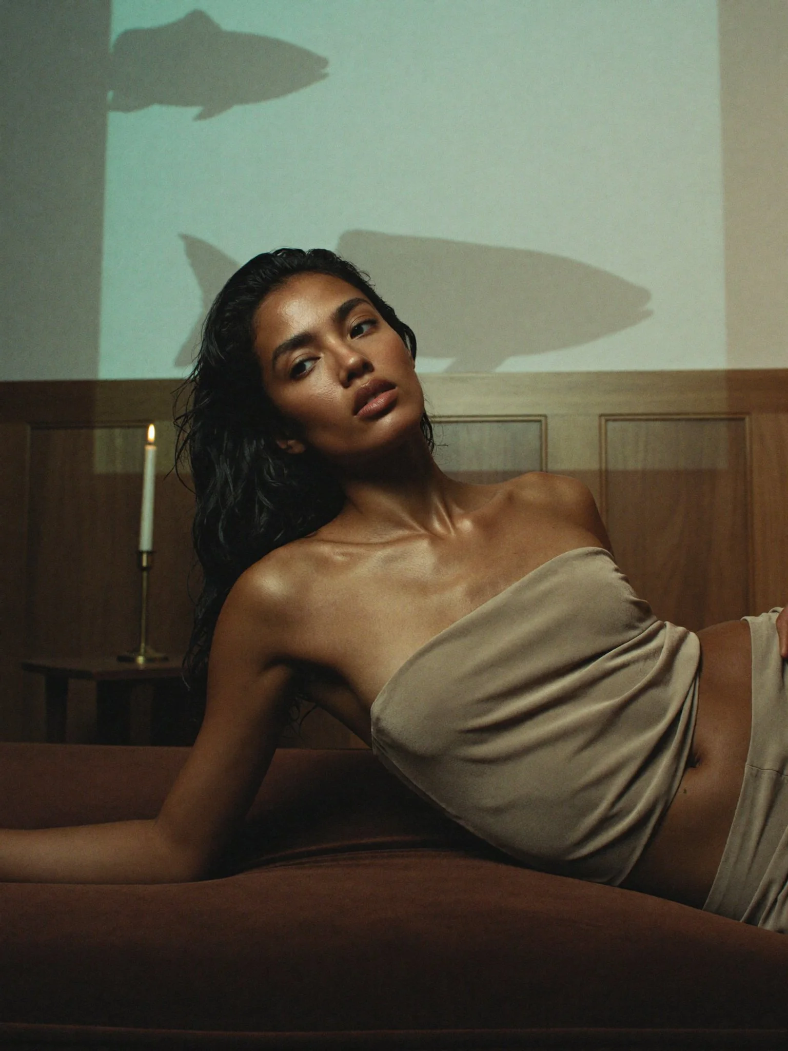 A woman with dark, wet hair and tan skin resting against a wooden headboard, wearing a silky beige off-shoulder top, with shadows of fish on the wall behind her, illuminated by a candle on a bedside table.