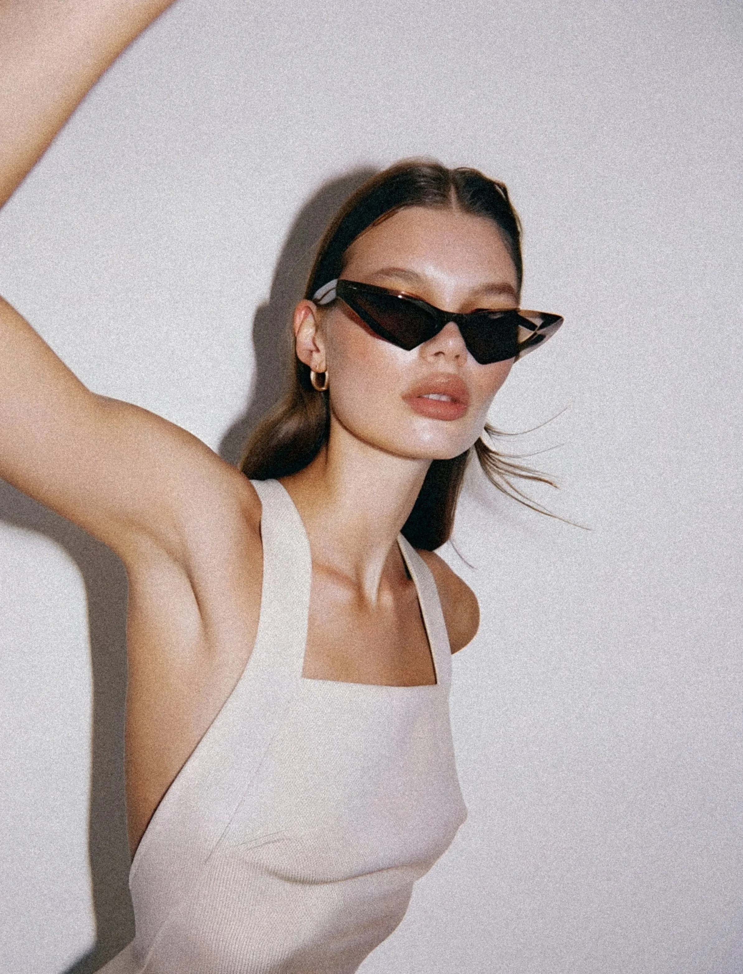 A woman with long dark hair wearing black sunglasses and gold earrings, dressed in a white tank top, posing against a textured white wall.