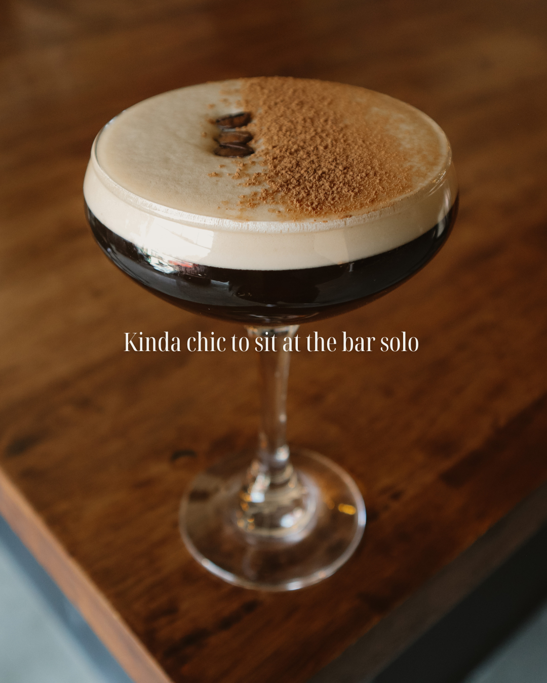 A cocktail in a coupe glass with a creamy foam top, coffee beans, and cocoa powder garnish, sitting on a wooden surface.