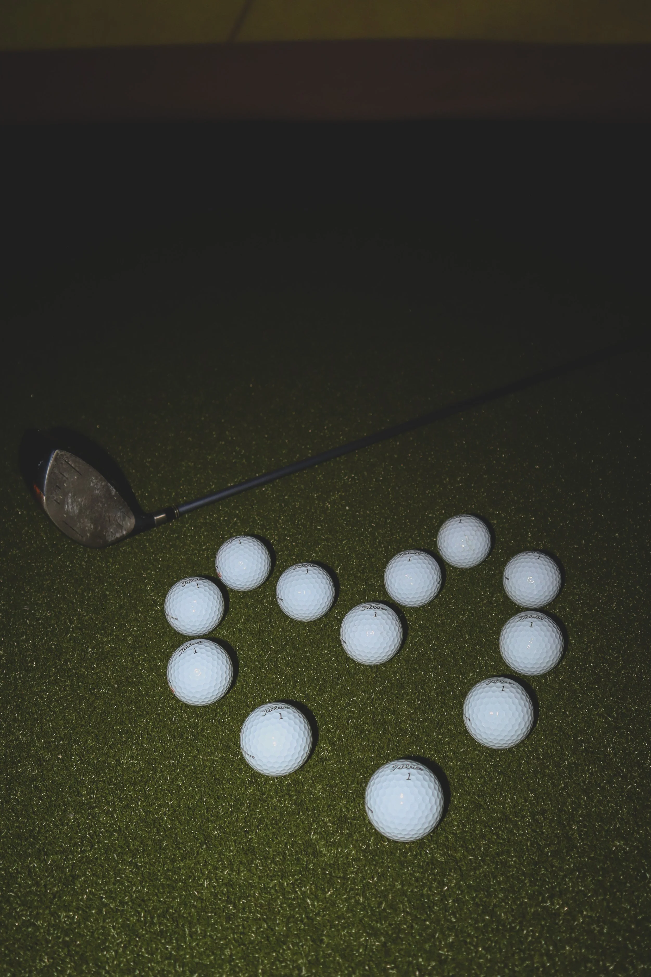 Golf club and multiple golf balls arranged on a green artificial turf, with a dark background.