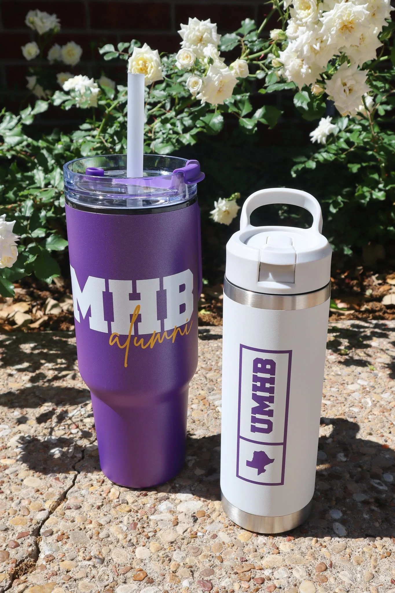 Two reusable drink containers, one purple with a straw and the word 'MICH' visible, and one white with a handle, placed on gravel in front of white roses.