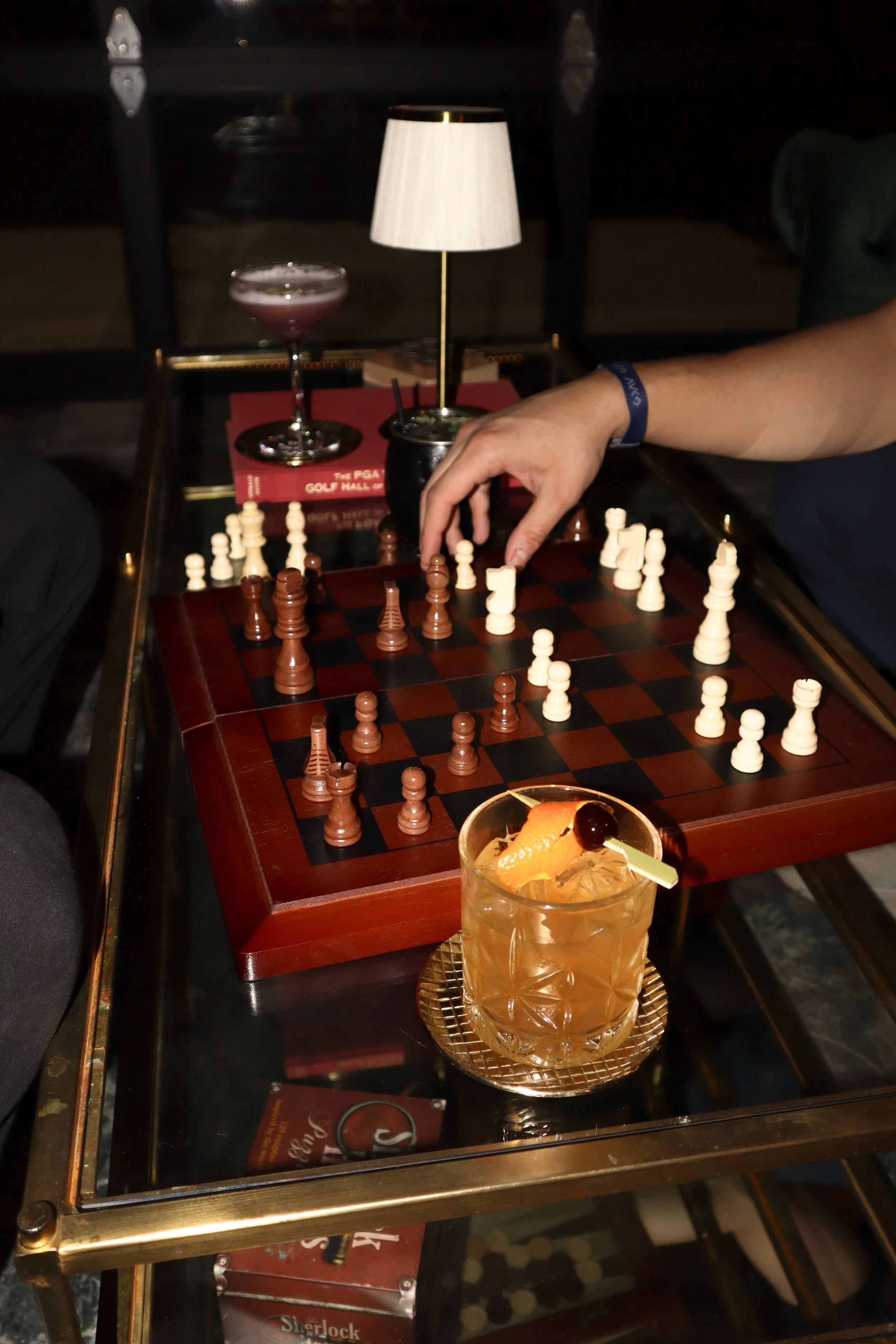A person playing chess at a table with a lamp, a cocktail on a tray, and a book titled 'The PGA Golf Hall of Fame' in the background.