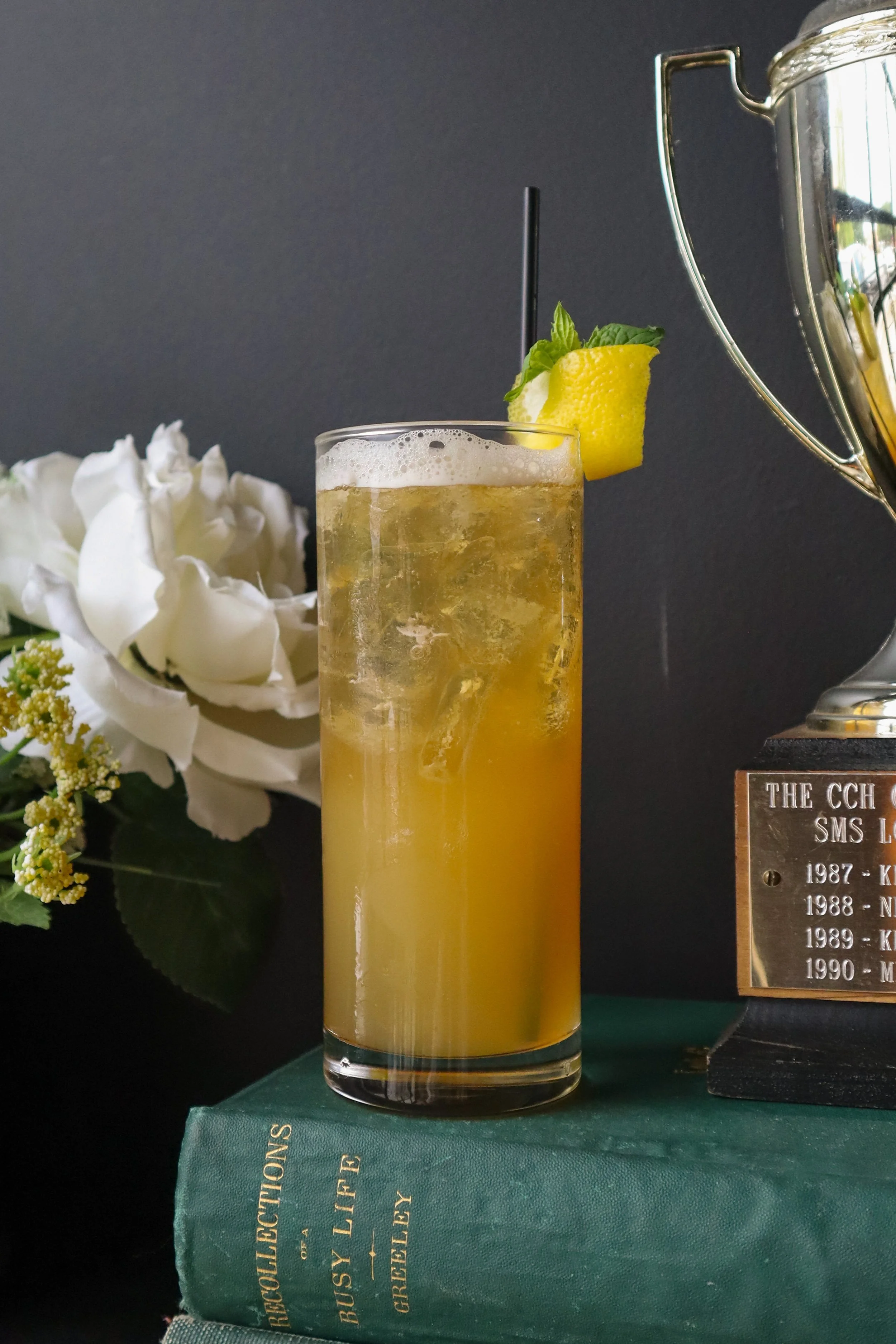 A tall glass of a yellow-orange beverage garnished with a lemon wedge and a sprig of mint, with a black straw, sitting on a green book with a bouquet of white flowers in the background.