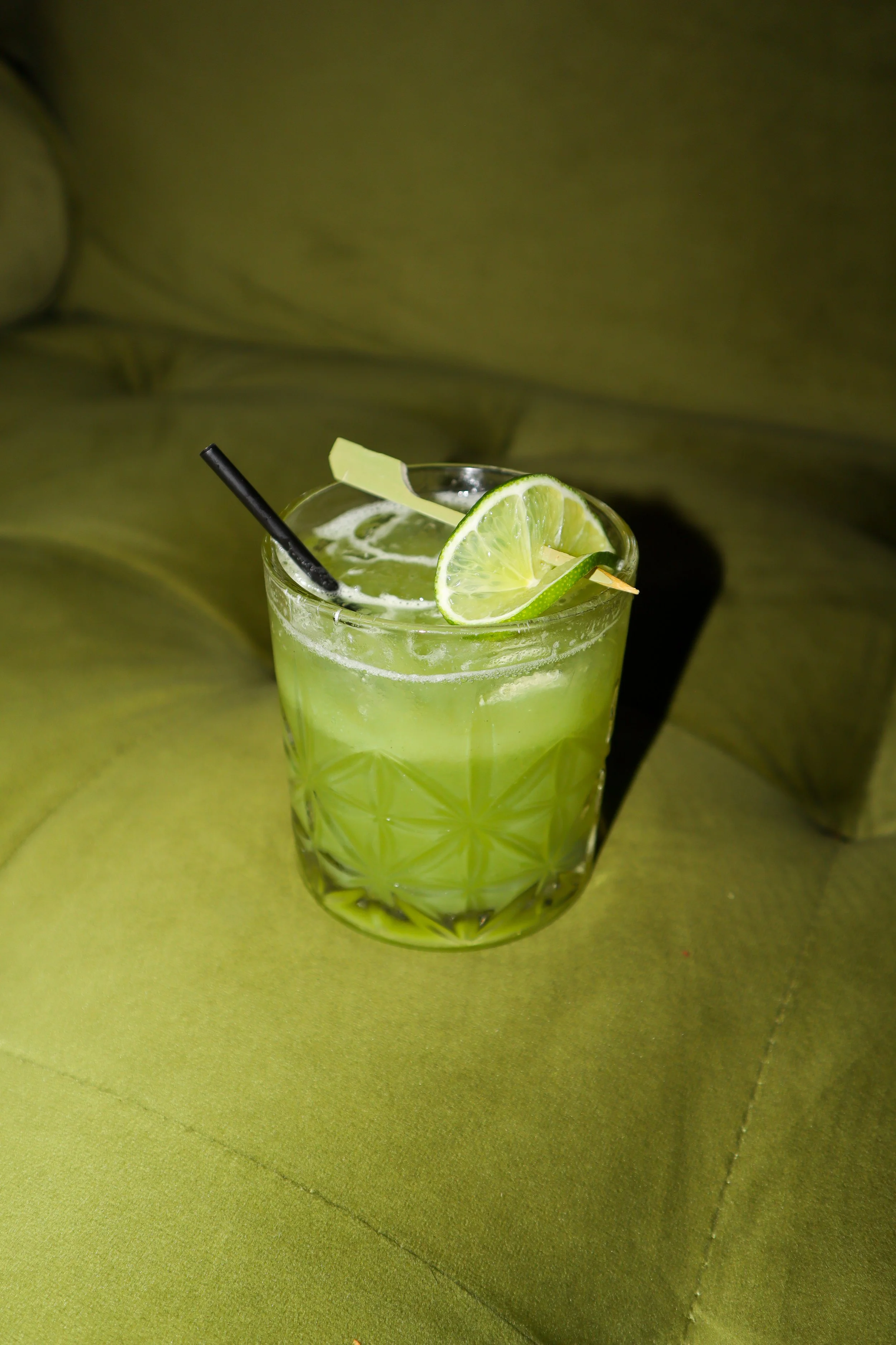 A glass of green beverage garnished with lime slices and a straw, set against a green textured background.