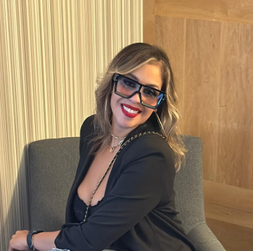 A woman with wavy blonde hair wearing glasses and a black blazer, smiling and sitting on a gray chair indoors.