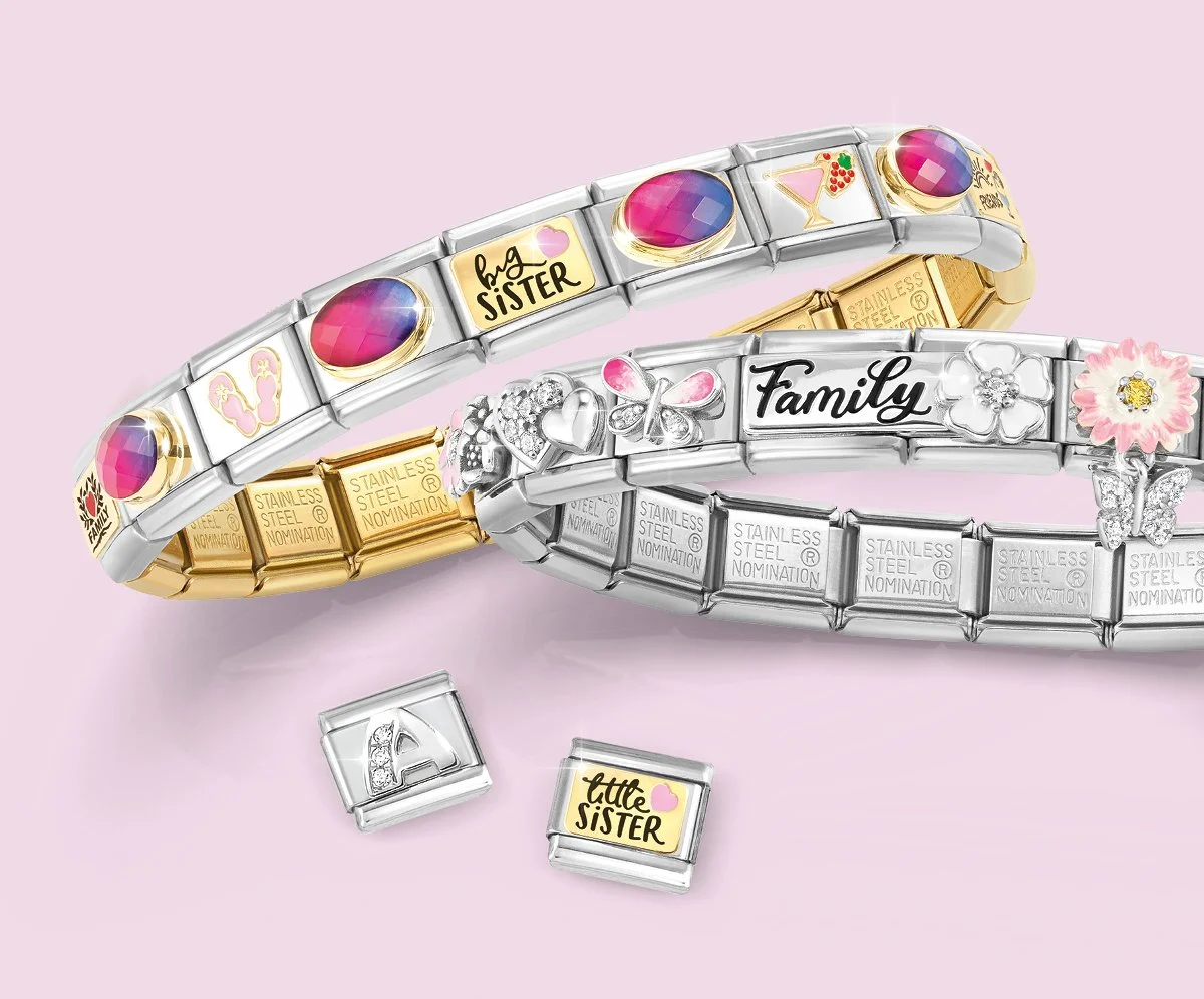 Three decorative Nomination Italy Composable Charm metal bracelets with colorful charms and embellishments, lying on a pastel pink background, with two small charms placed beneath them.