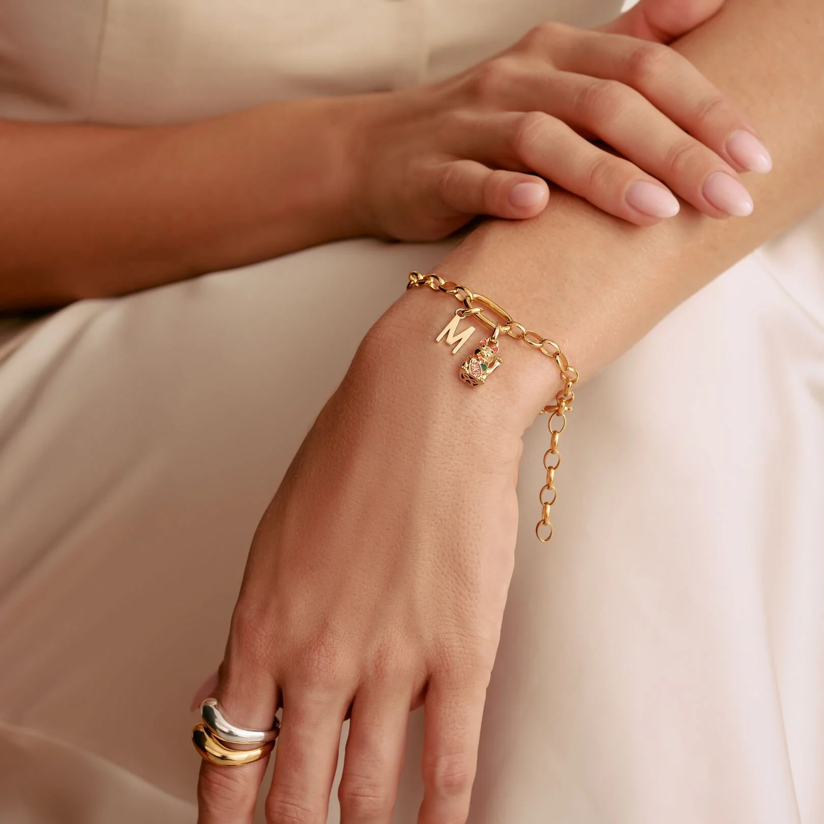 Close-up of a woman's hand and wrist wearing gold Thomas Sabo jewelry, including a bracelet with letter 'M', a charm with a bear, and a multi-colored ring, against light-colored fabric.