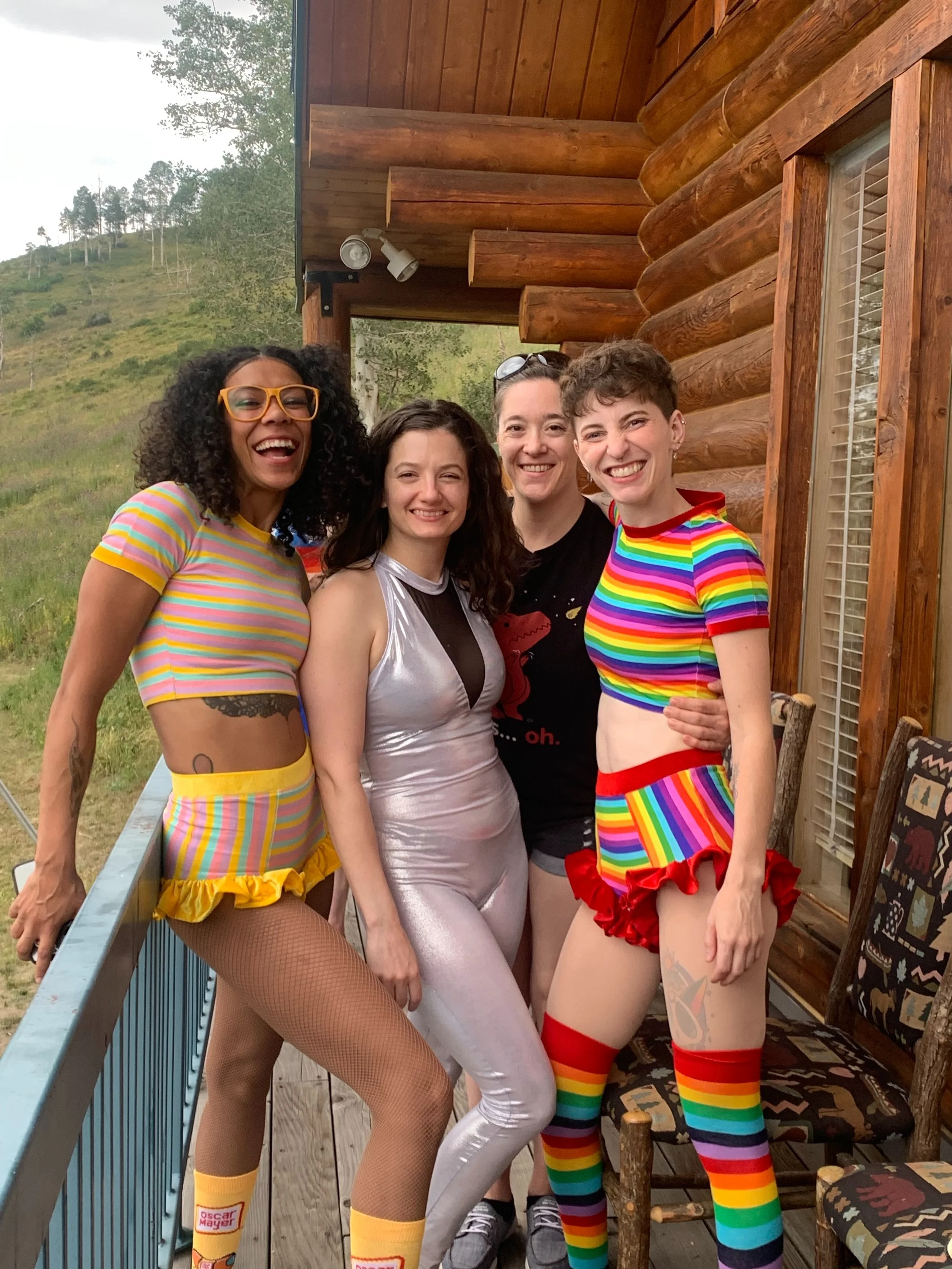Four women in colorful, rainbow-themed outfits smiling on a porch with a wooden log cabin in the background and a scenic hillside.
