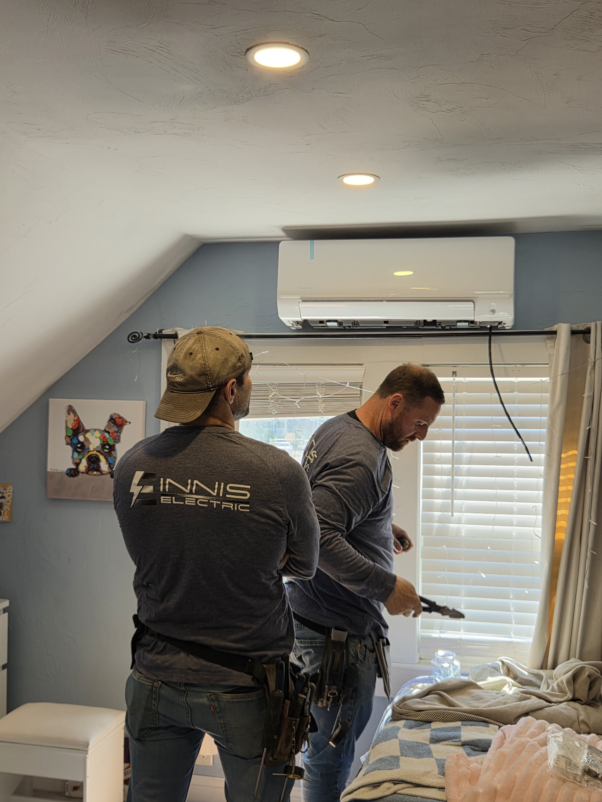 Two men working together in a bedroom, one holding tools, near a bed with a blanket, a window with blinds, and an air conditioning unit on the wall.