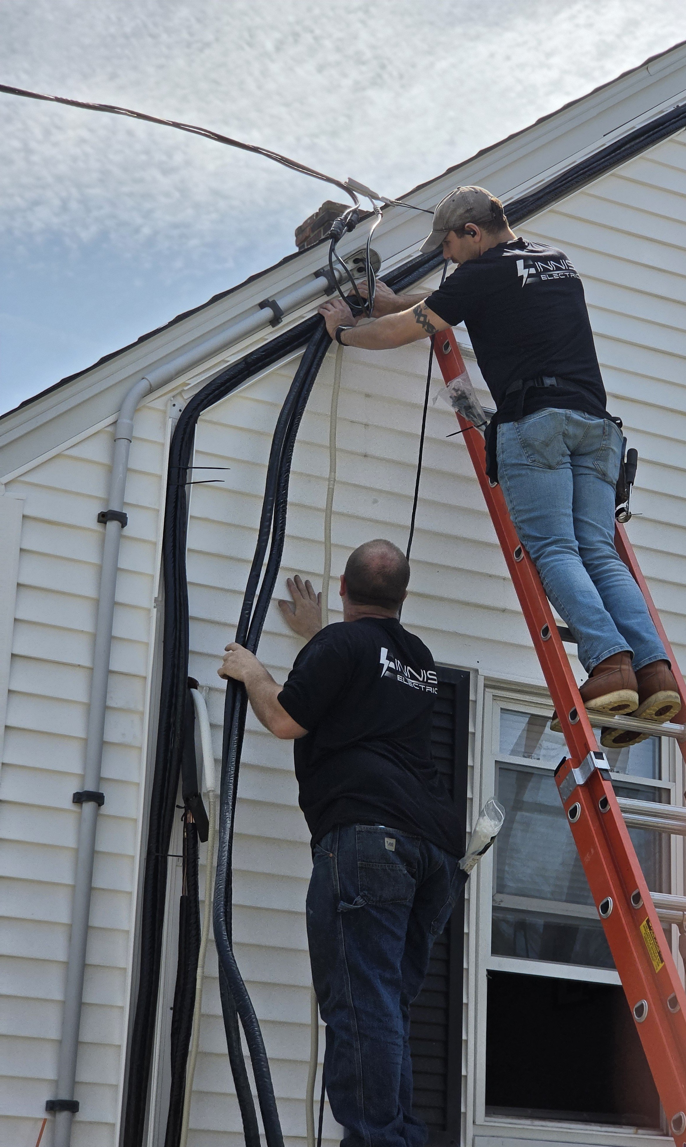 Two technicians installing or inspecting electrical wires on the side of a beige house, one standing on a ladder and the other on the ground.