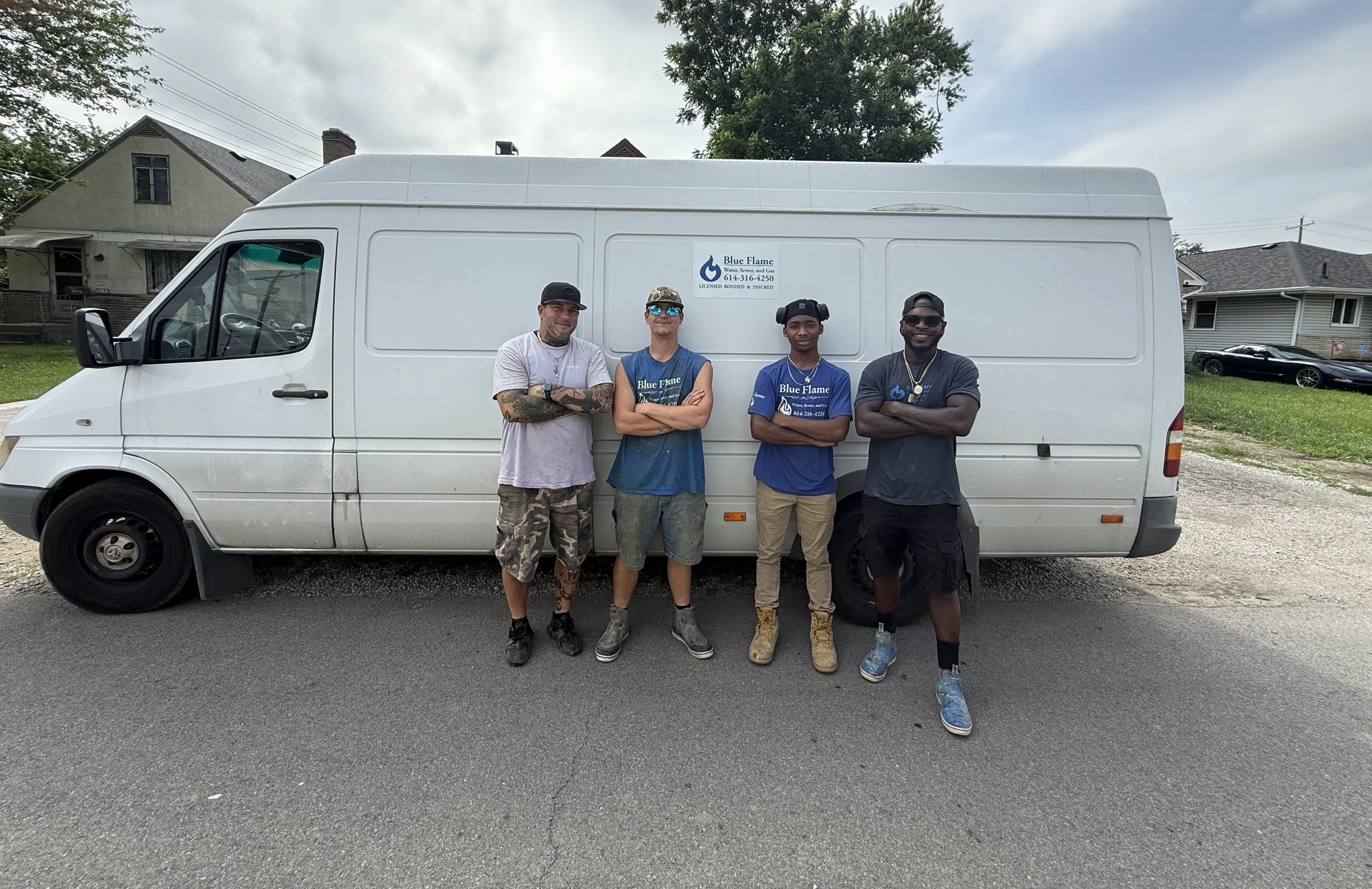 Four men standing with crossed arms in front of a white service van with a logo for Blue Flame on its side, parked on a street with houses and trees in the background.