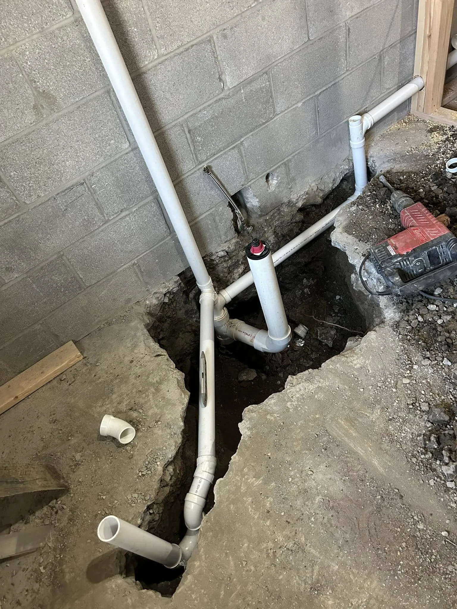 Under construction plumbing pipes installed in a trench in a basement or utility room with a concrete block wall in the background.