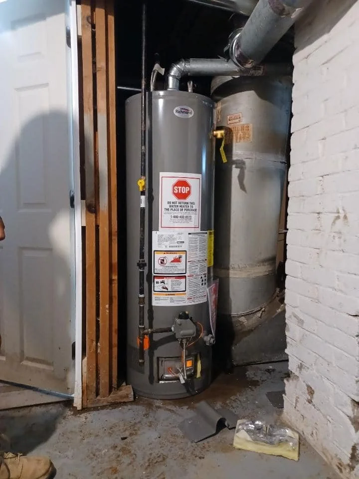 A gray Rheem water heater installed next to a large duct in a basement or utility room, with warning labels and pipes connected at the top.