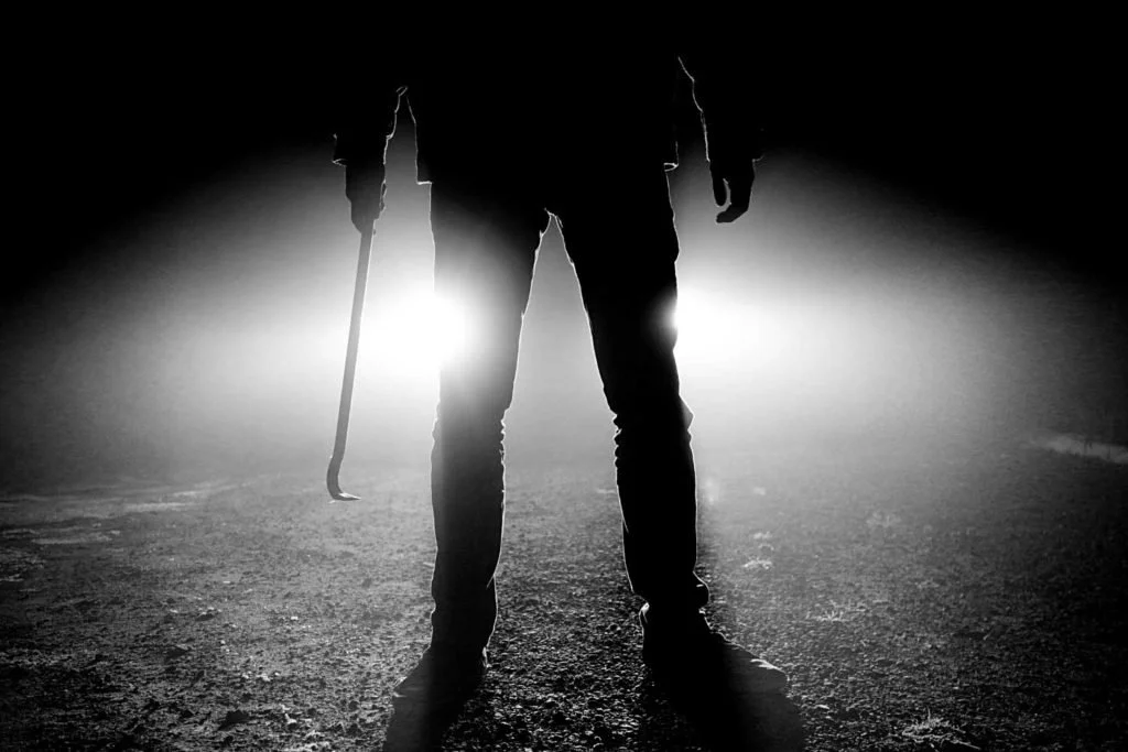 Silhouette of a person holding a cane, backlit by bright lights, on a dark outdoor road at night.