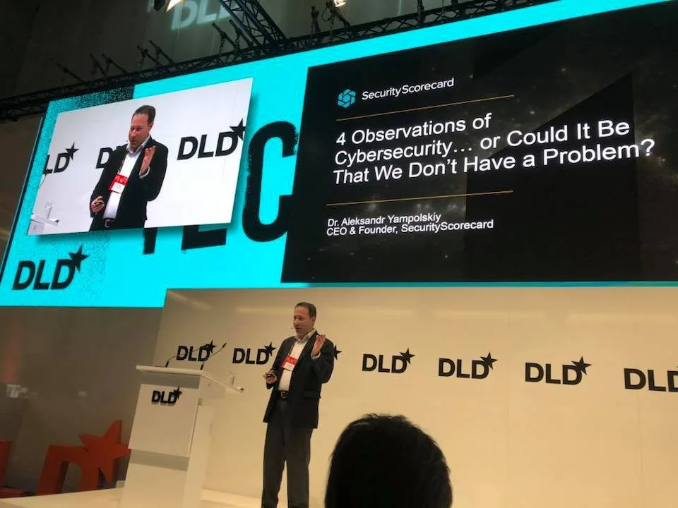 Speaker at a DLD conference giving a presentation on cybersecurity, titled "4 Observations of Cybersecurity... or Could It Be That We Don’t Have a Problem?" by Dr. Aleksandr Yampolskiy, CEO and Founder of SecurityScorecard.