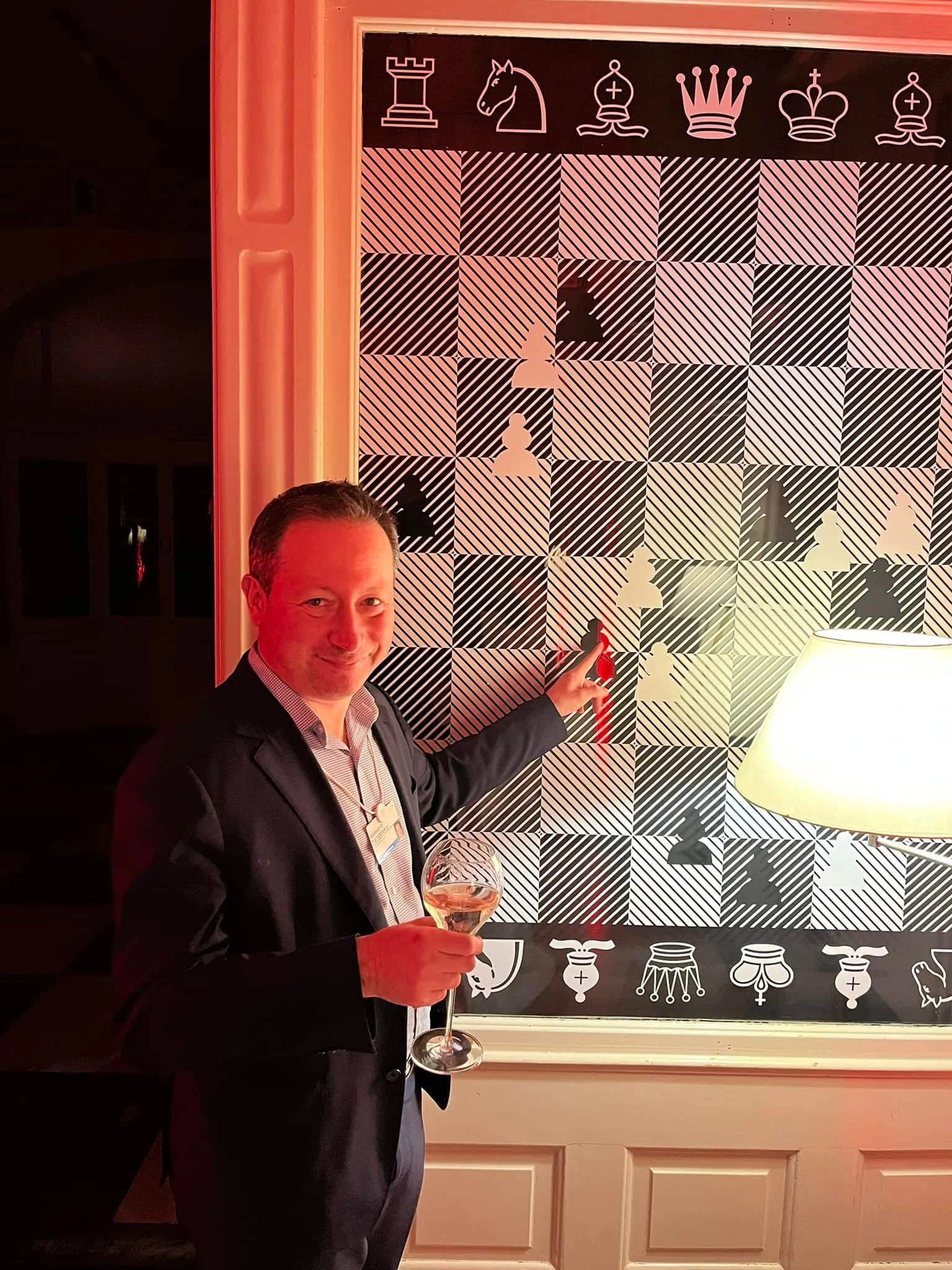 Dr. Aleksandr Yampolskiy | Cybersecurity Expert & CEO of Security in a suit holding a glass of wine, pointing at a chessboard-themed artwork with chess pieces silhouettes and decorative symbols on the top and bottom, in a dimly lit room with a lamp.
