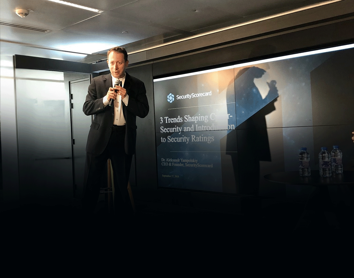 Dr. Aleksandr Yampolskiy | Cybersecurity Expert & CEO of SecurityScorecard in a black suit and white shirt speaking into a microphone in front of a presentation screen at a conference.