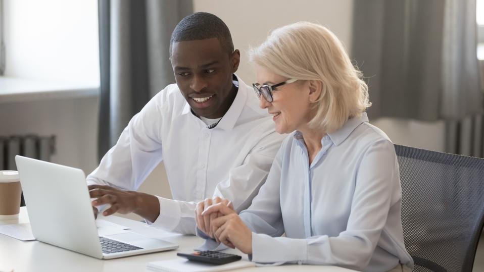 A young man and an older woman sit at a desk, looking at a laptop screen and smiling, in a well-lit office.