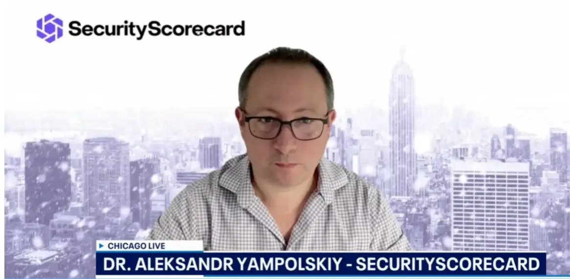A man wearing glasses and a checkered shirt appears on a news broadcast, with the SecurityScorecard logo in the top left corner and a city skyline background. A news ticker at the bottom reads 'CHICAGO LIVE' and 'DR. ALEKSANDR YAMPOLSKIY - SECURITYSCORECARD'.