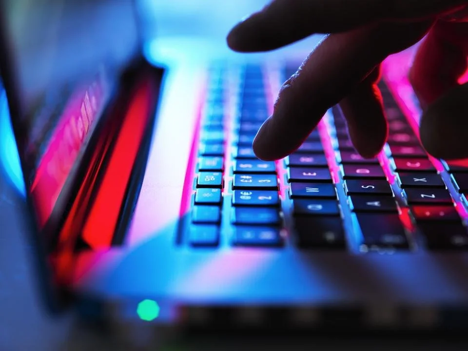 Close-up of a person's hand typing on a backlit keyboard with pink and blue lights.