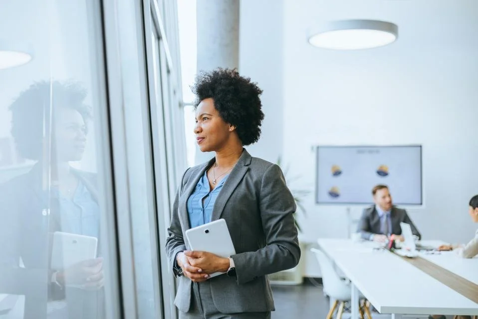 Businesswoman standing by window in a modern conference room, holding a tablet, with colleagues seated at a table in the background.
