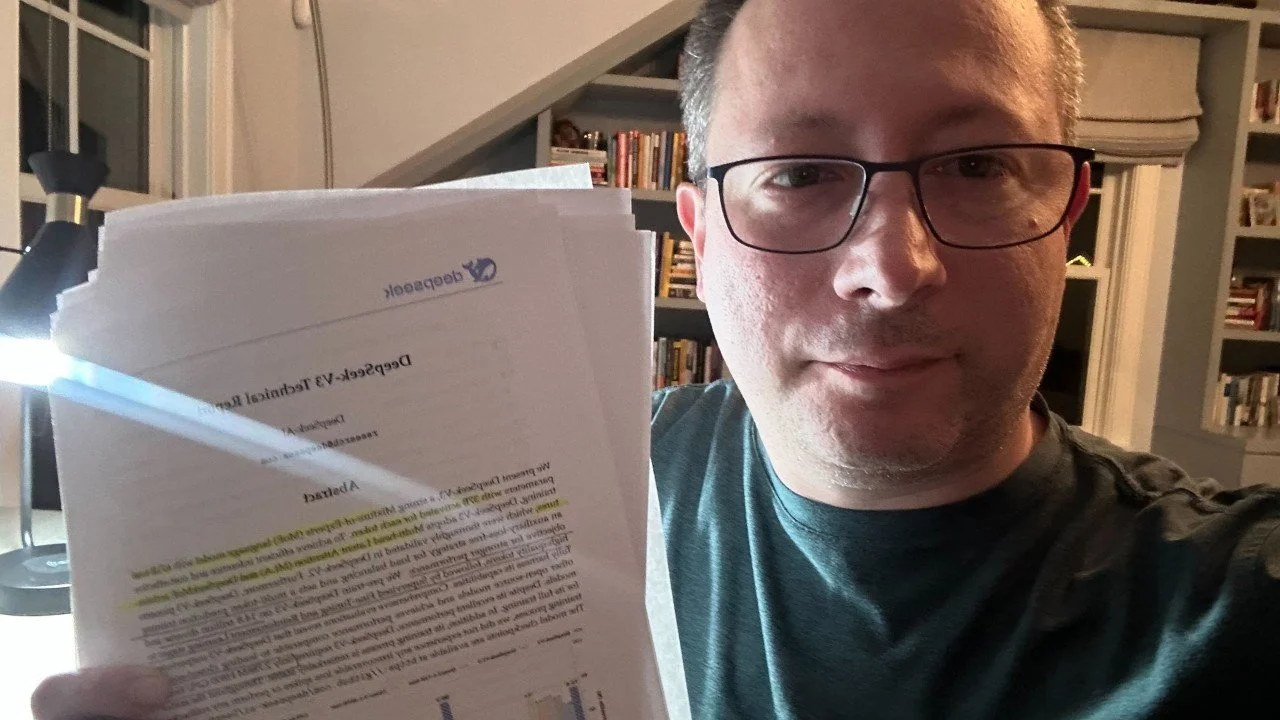 Dr. Aleksandr Yampolskiy | Cybersecurity Expert & CEO of SecurityScorecard wearing glasses holding a stack of papers in a room with bookshelves and a ceiling corner, illuminated by a desk lamp.