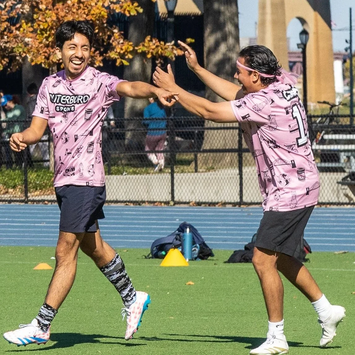 Two men celebrating on a sports field, wearing matching pink sports jerseys with printed designs, shorts, and sneakers. They are smiling and high-fiving each other.