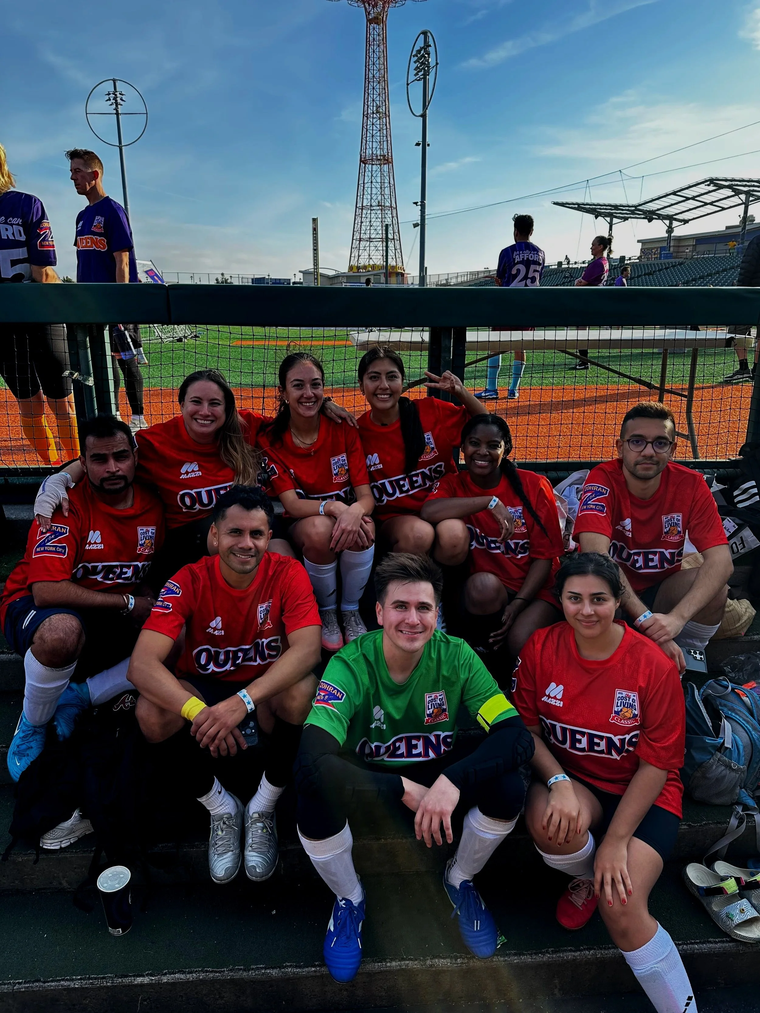 A group of nine young soccer players, six women and three men, sitting and kneeling on the bench in soccer uniforms, smiling at the camera with a soccer field and spectators in the background.