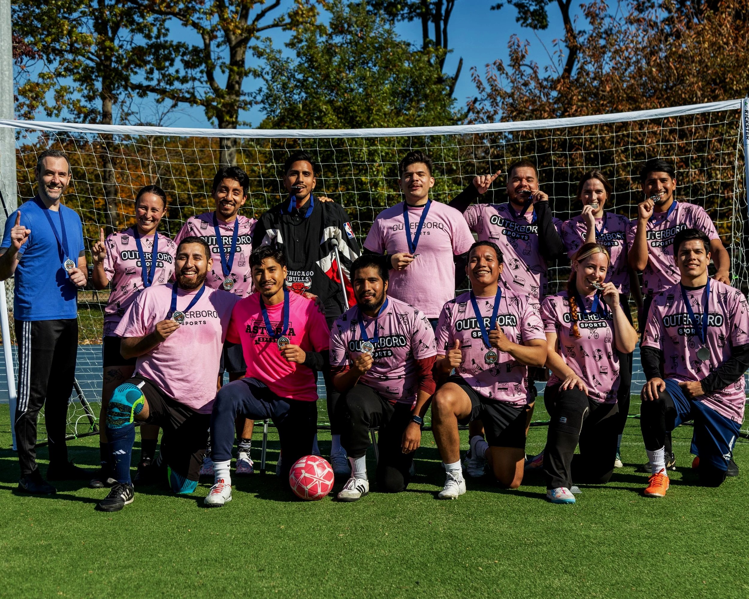 A women's soccer team and coaches celebrating on the field after winning a match, all wearing medals and posing in front of a goal on a sunny day.