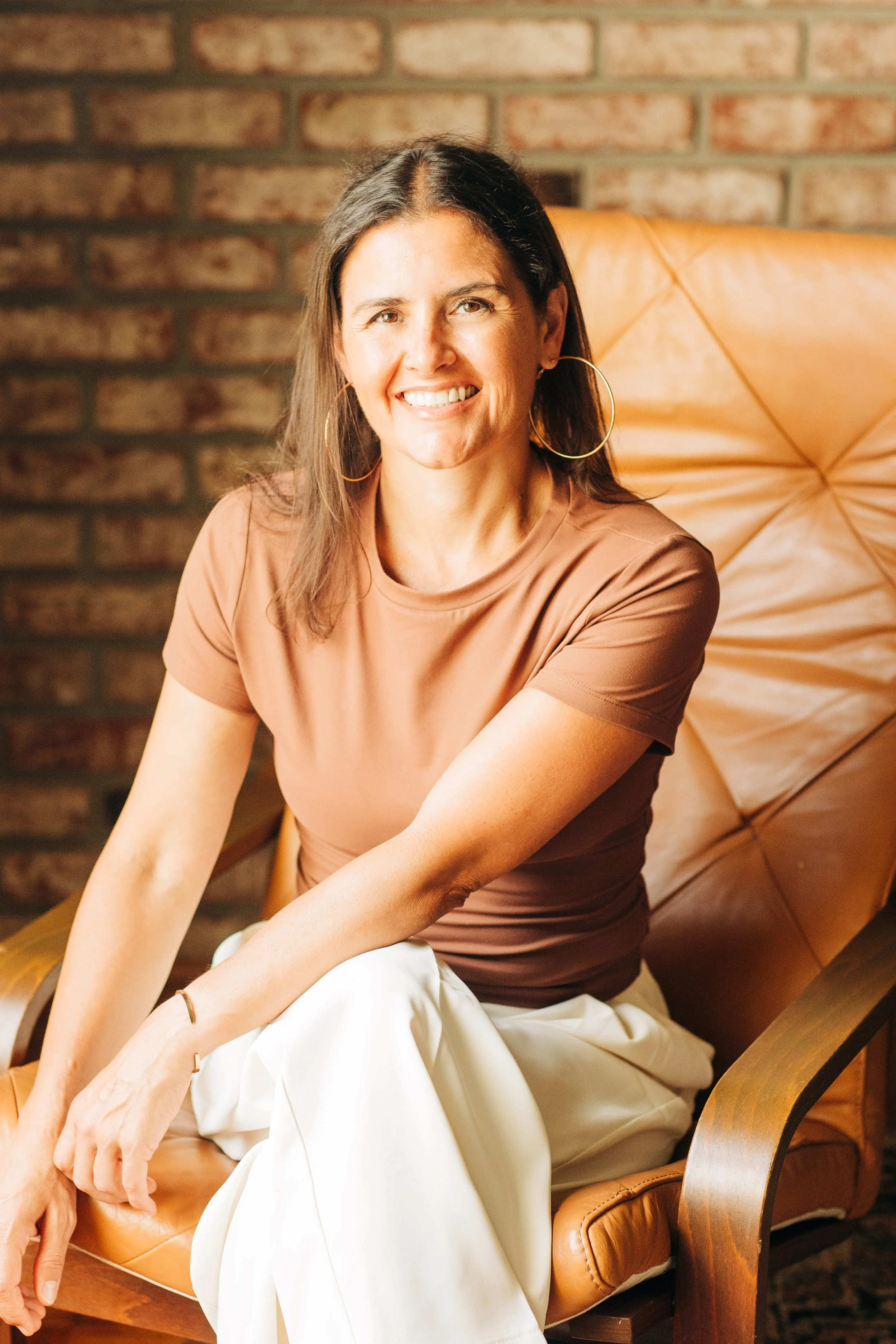 A woman with long brown hair sitting on a tan leather chair with a brick wall background, smiling and wearing a beige t-shirt and white pants.