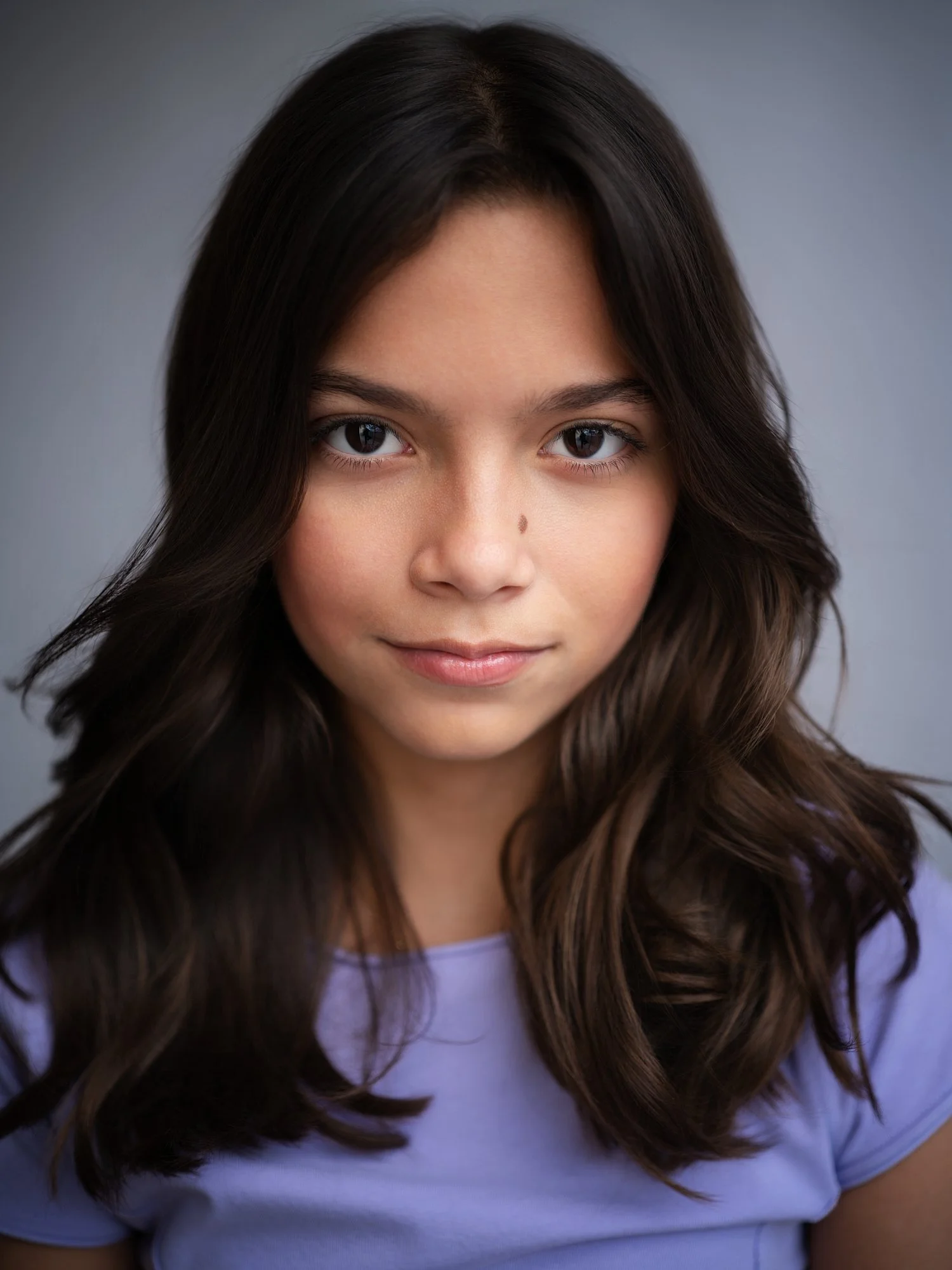 Close-up portrait of a young woman with long, wavy brown hair, wearing a light purple shirt, against a neutral background.