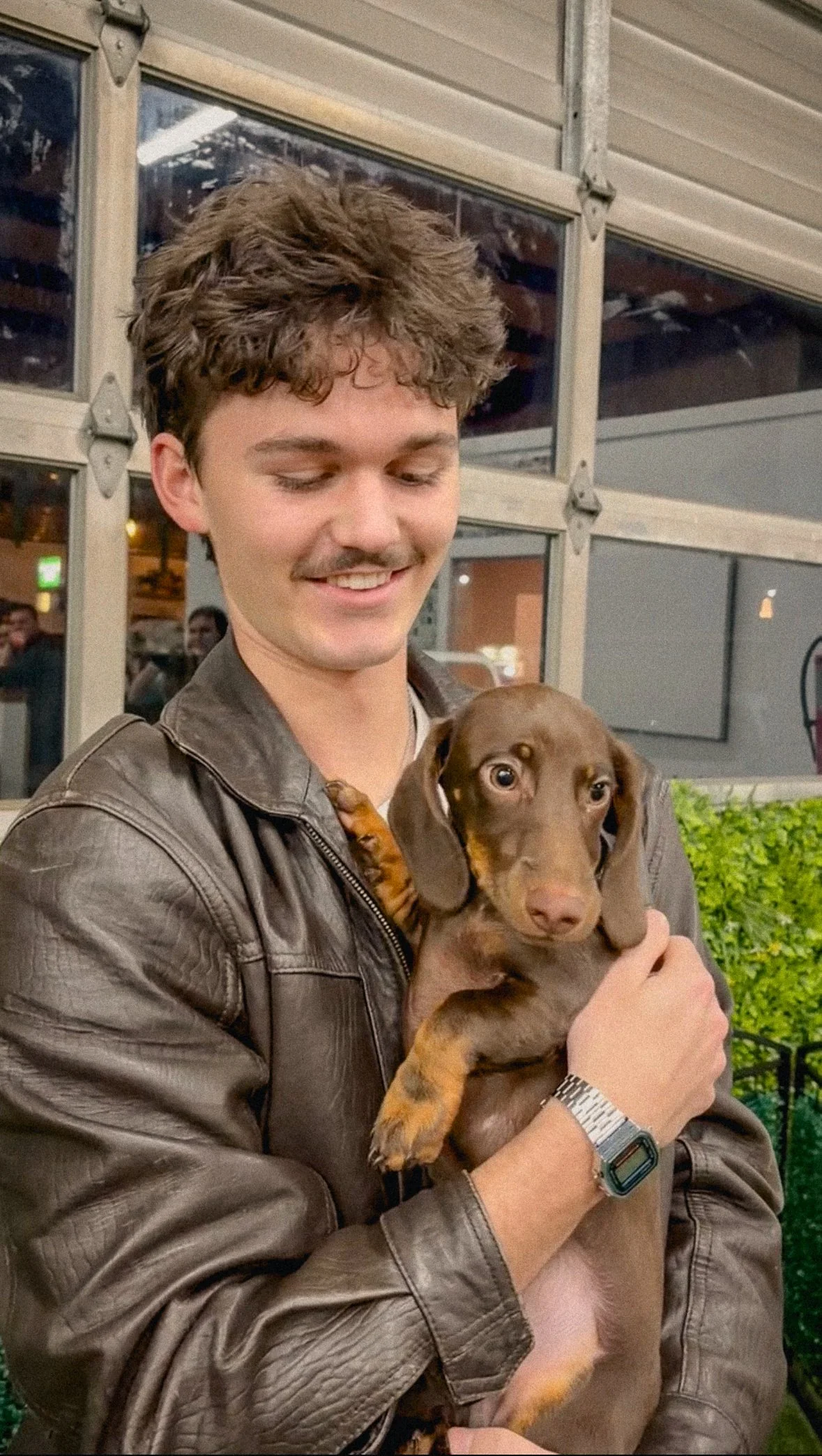 A young man with curly brown hair, wearing a brown leather jacket, is holding a small brown and tan dachshund puppy. They are indoors, with a background showing large glass windows and some people in the distance.