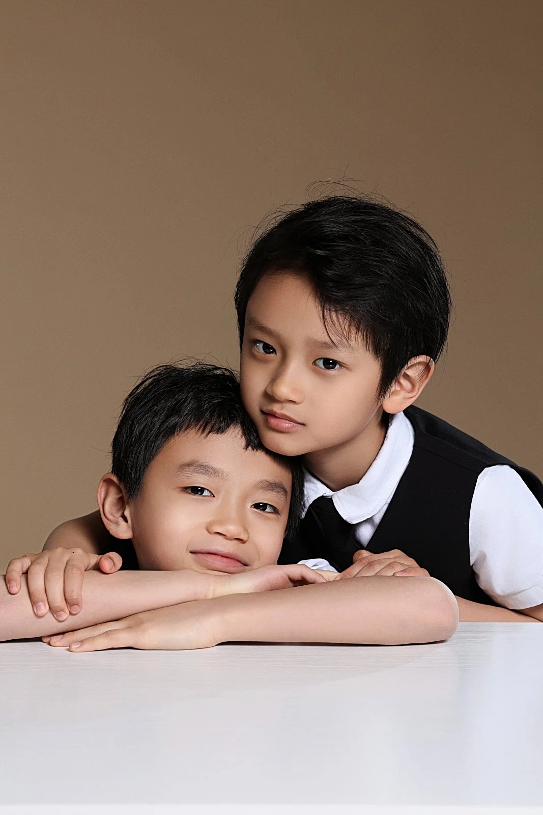 Two young boys with Asian features, one older and one younger, pose closely against a plain background. The older boy has black hair and is wearing a white shirt and black vest, while the younger boy has short black hair and is resting his head on hi
