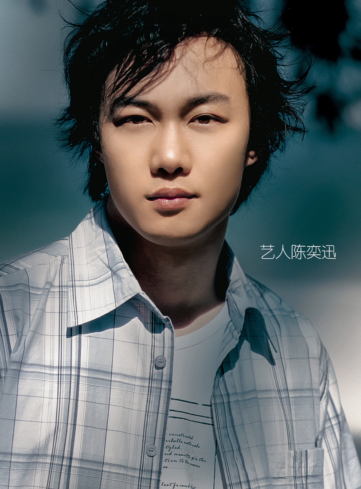 A young man with dark, slightly messy hair outdoors on a sunny day, wearing a light plaid shirt over a white graphic T-shirt, with a blurred background of trees and sky.
