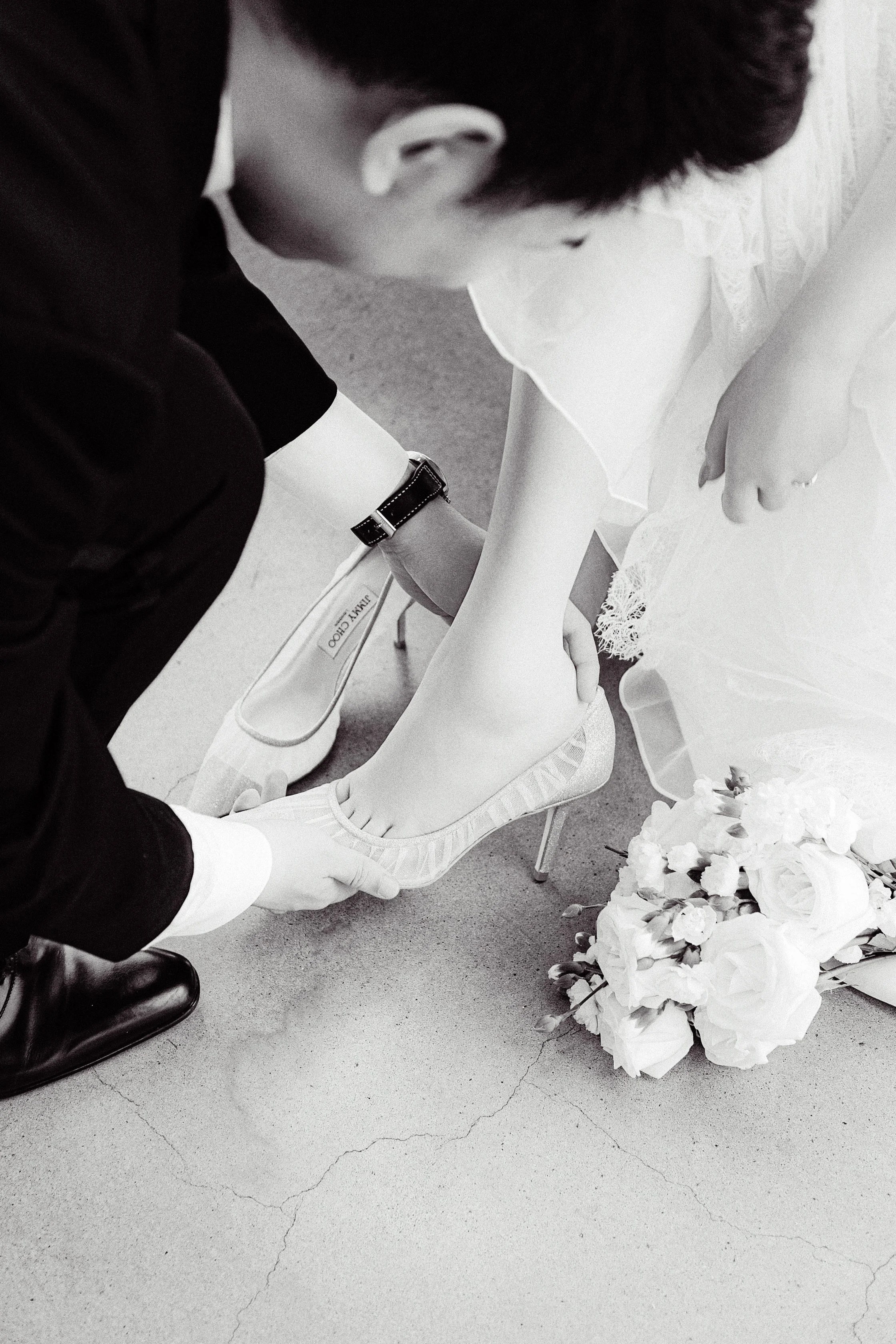 A man in a tuxedo and a woman in a wedding dress are holding hands, with the woman sitting on a chair and the man kneeling in front of her. A bouquet of white flowers is on the floor nearby.