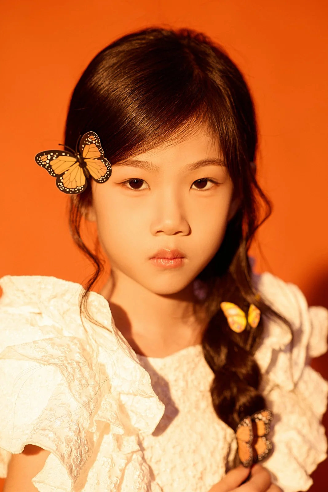 A young woman with long dark hair styled in a braid, wearing a white ruffled top, with three orange and black butterflies on her hair, shoulder, and holding in her hand, against a solid orange background.