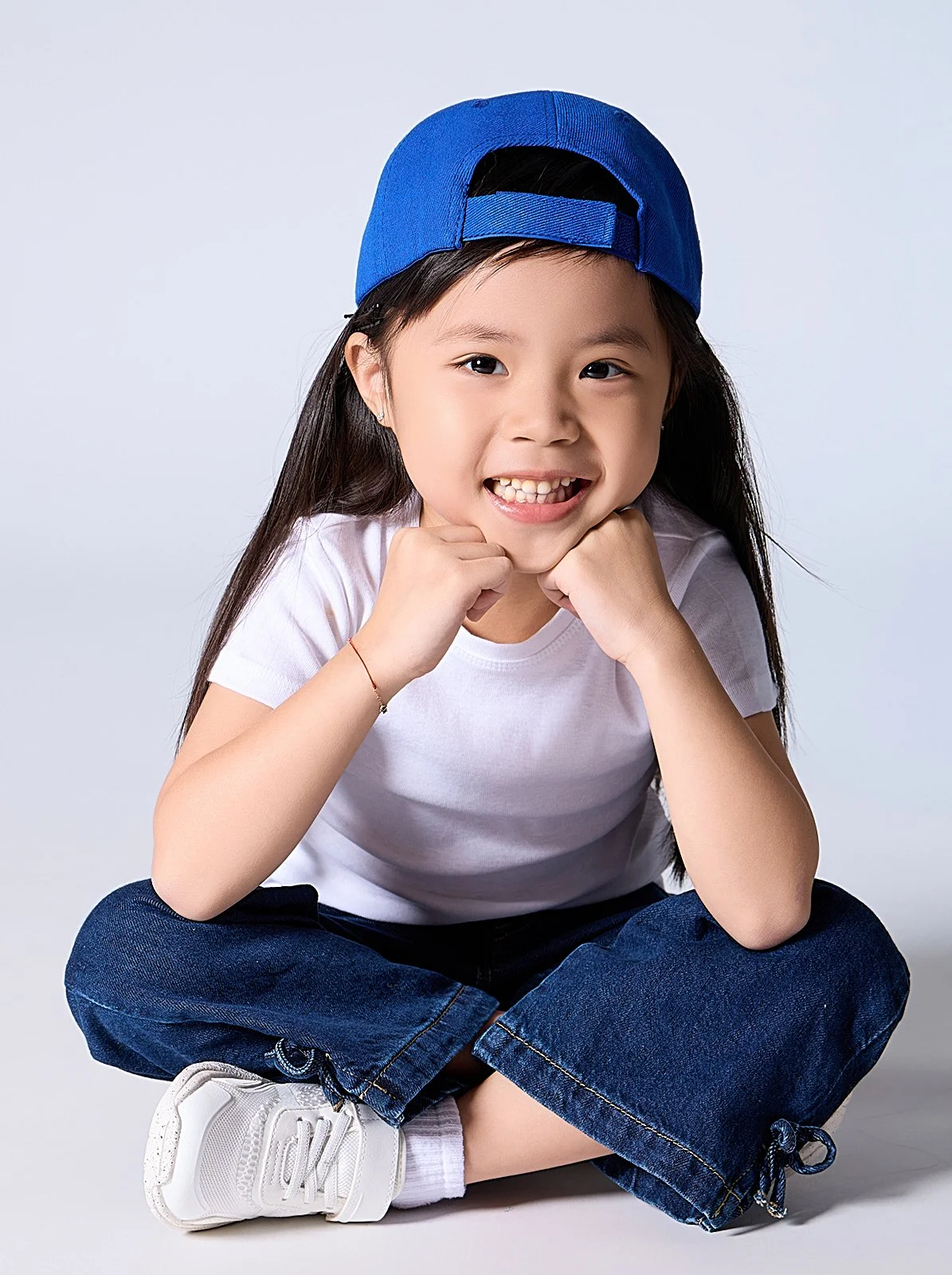Young girl with long dark hair, wearing a blue baseball cap backwards, white t-shirt, blue jeans, and white sneakers, sitting cross-legged and smiling with her hands under her chin against a plain background.