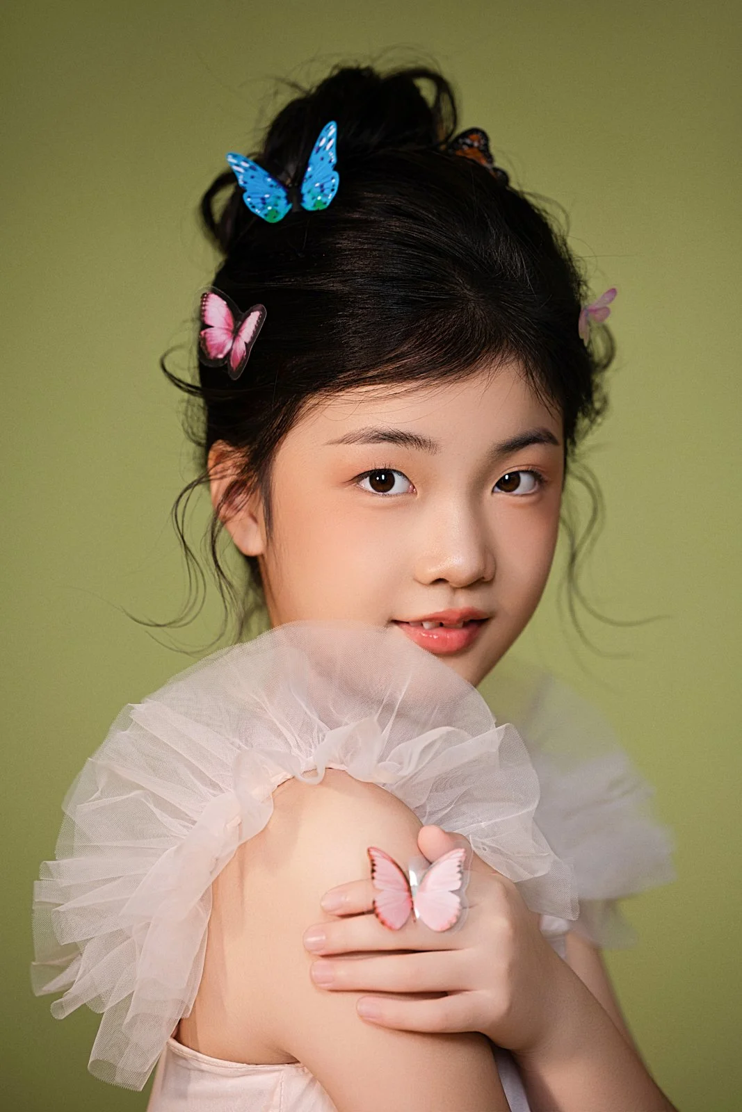 A young woman with dark hair styled in an updo decorated with colorful butterfly hair clips, holding a butterfly on her shoulder against a green background.