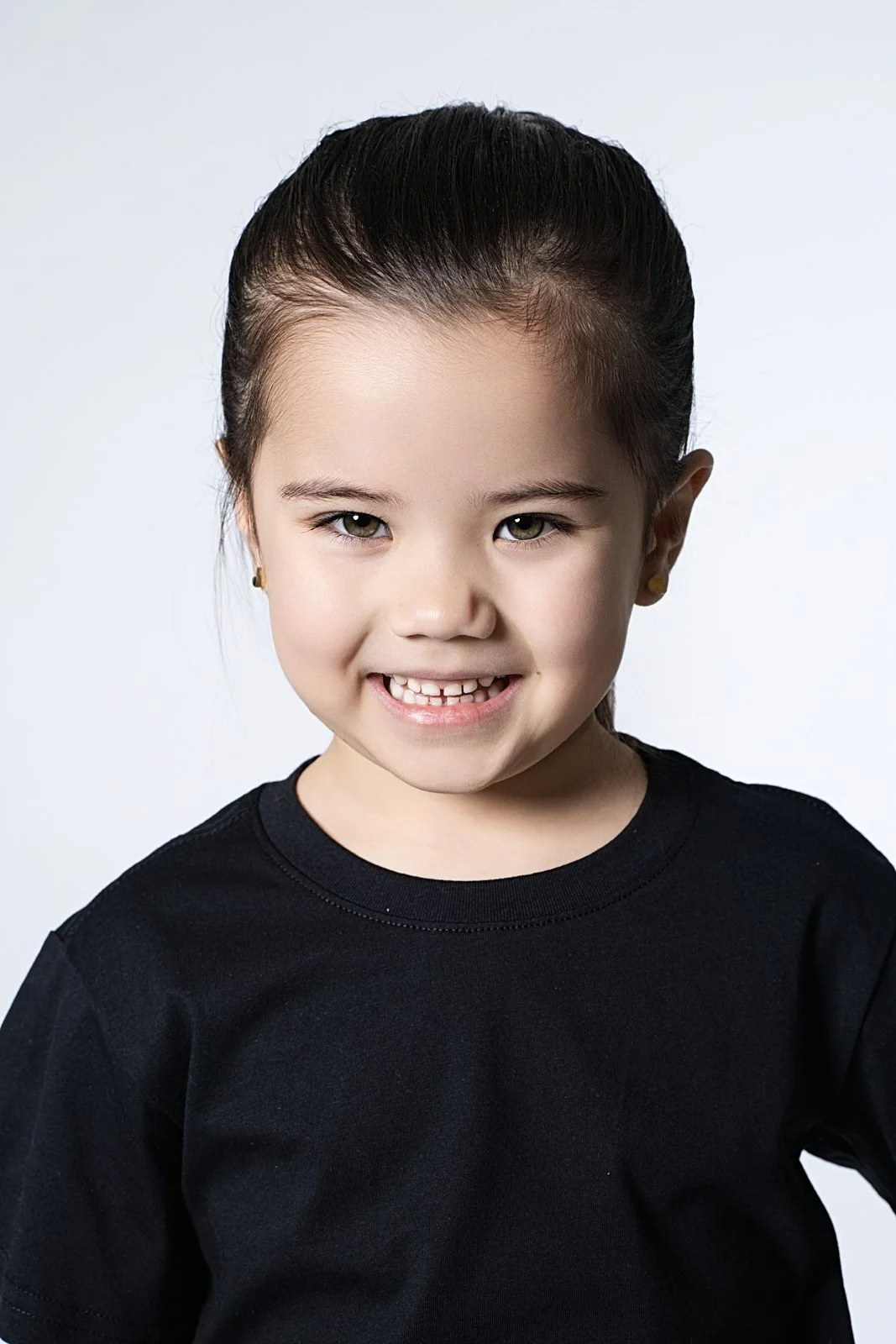 A young girl with dark hair tied back, wearing a black shirt, smiling with one missing front tooth, and looking at the camera against a light background.