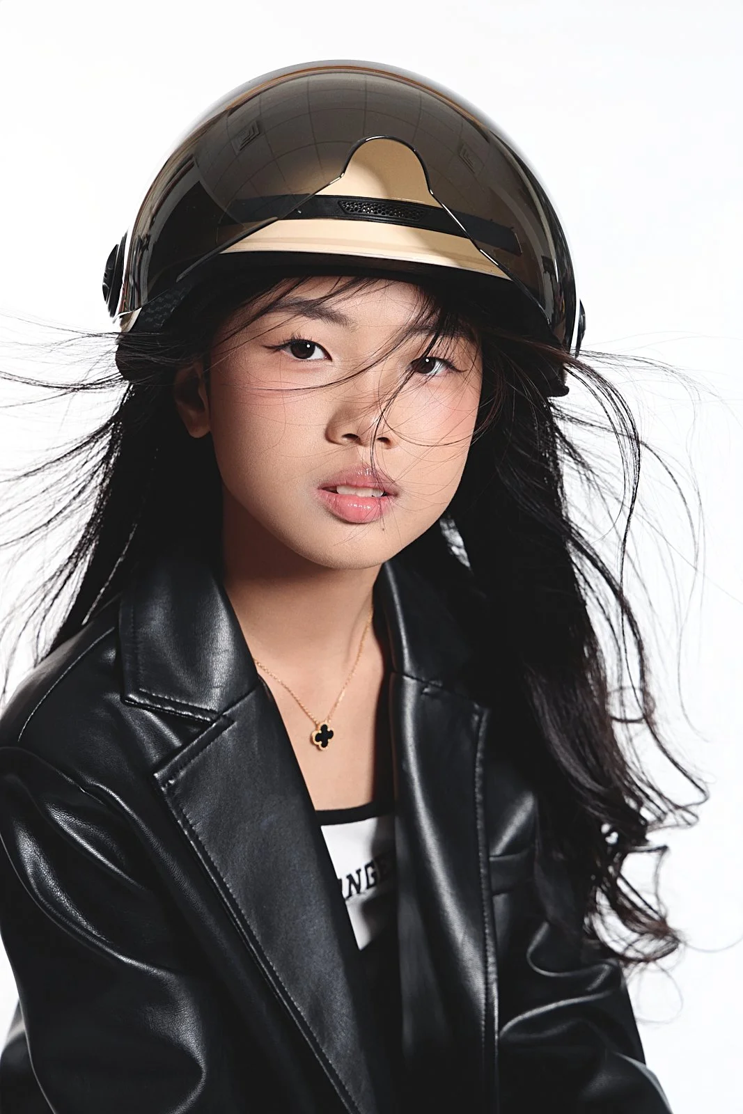 A woman with long black hair wearing a black motorcycle helmet, black leather jacket, and black and white shirt, posing against a white background.