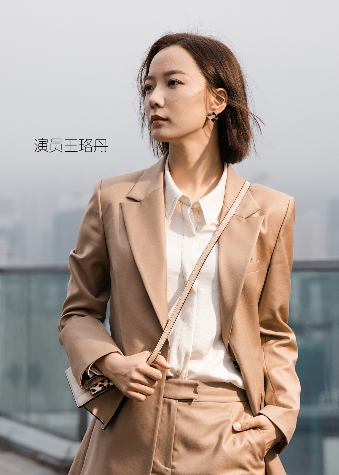 A young woman with shoulder-length brown hair standing outdoors in a cityscape, dressed in a beige blazer and matching pants, holding a purse with a leopard-print detail, with a serious expression.
