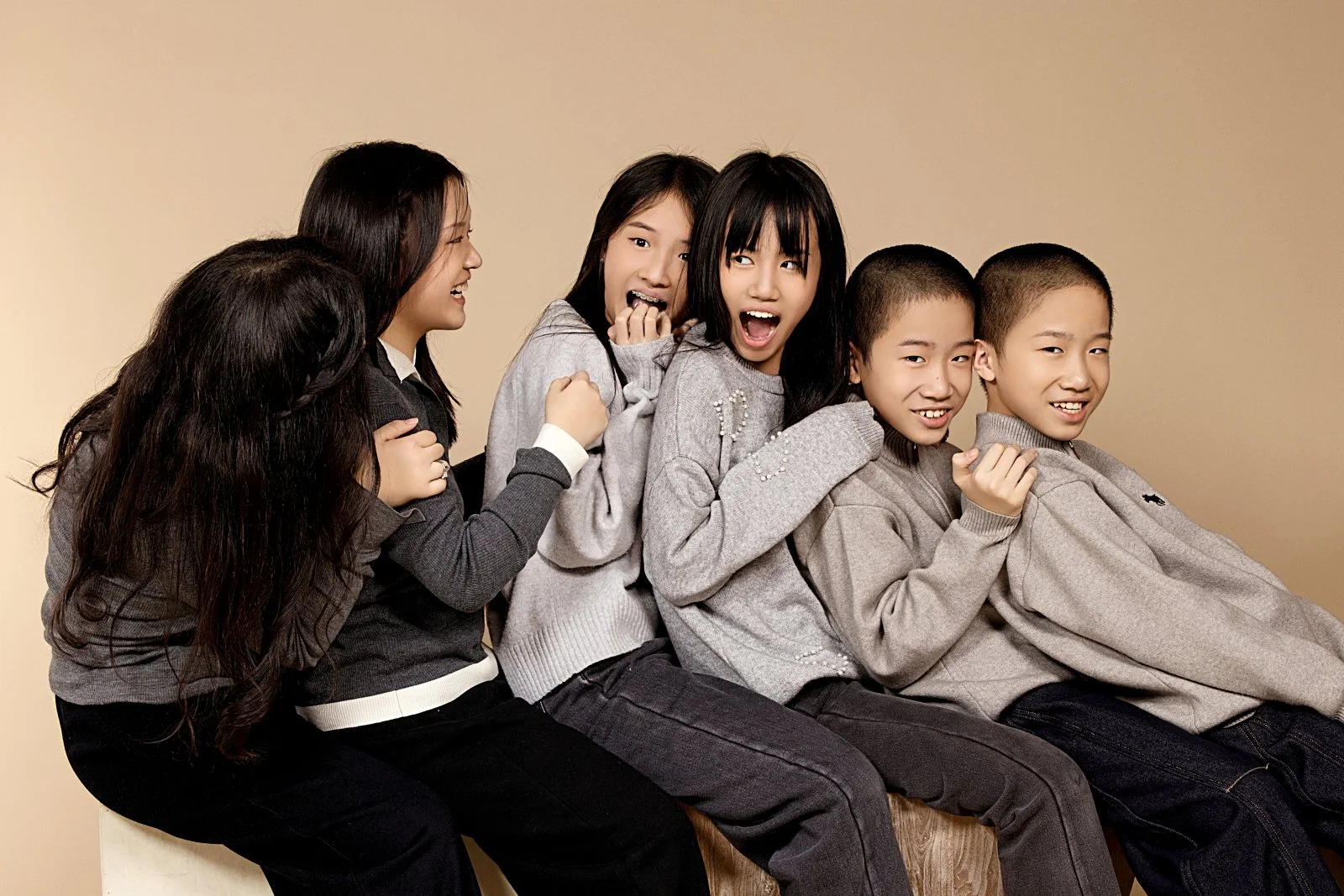 Six children sitting in a row, laughing and smiling, wearing casual clothes, against a plain beige background.