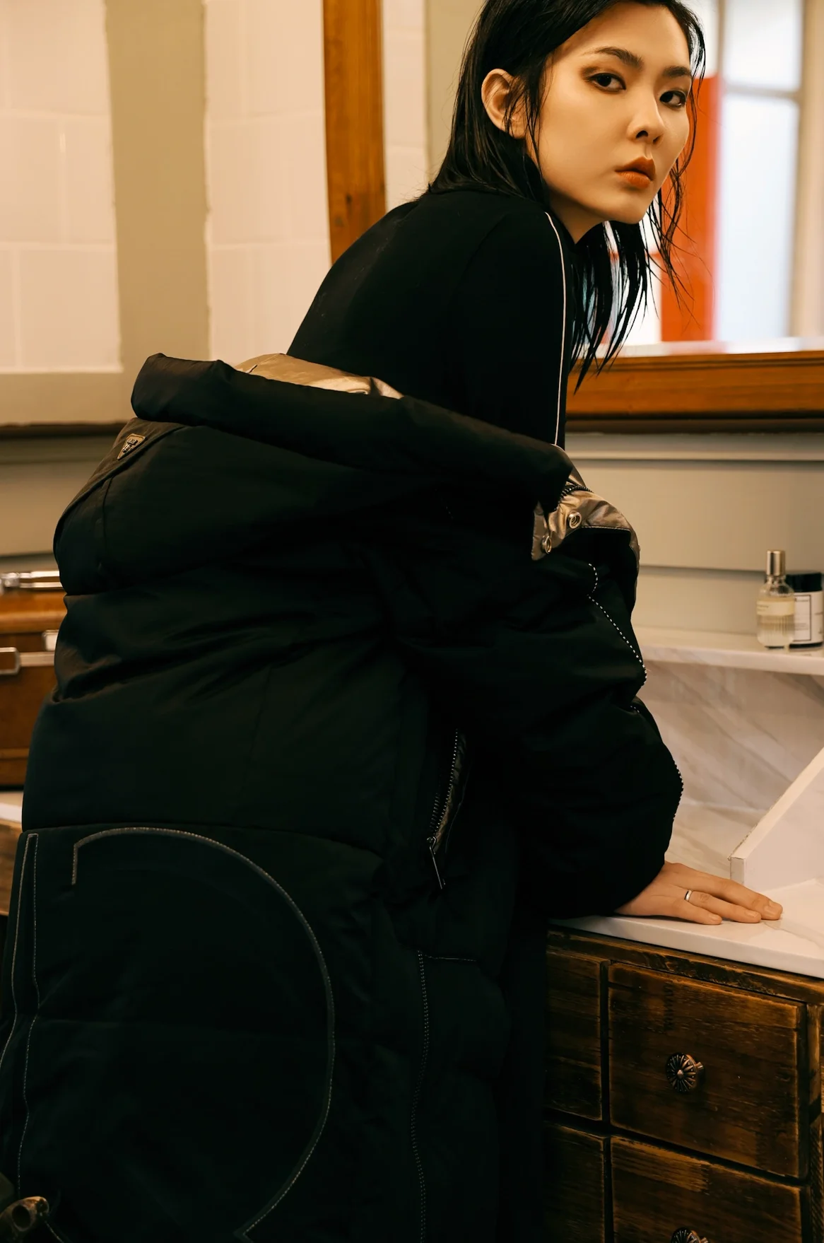 A woman with black hair and wearing a black jacket, leaning on a bathroom counter with her hand resting on the sink, in a room with wooden and white tiled walls.