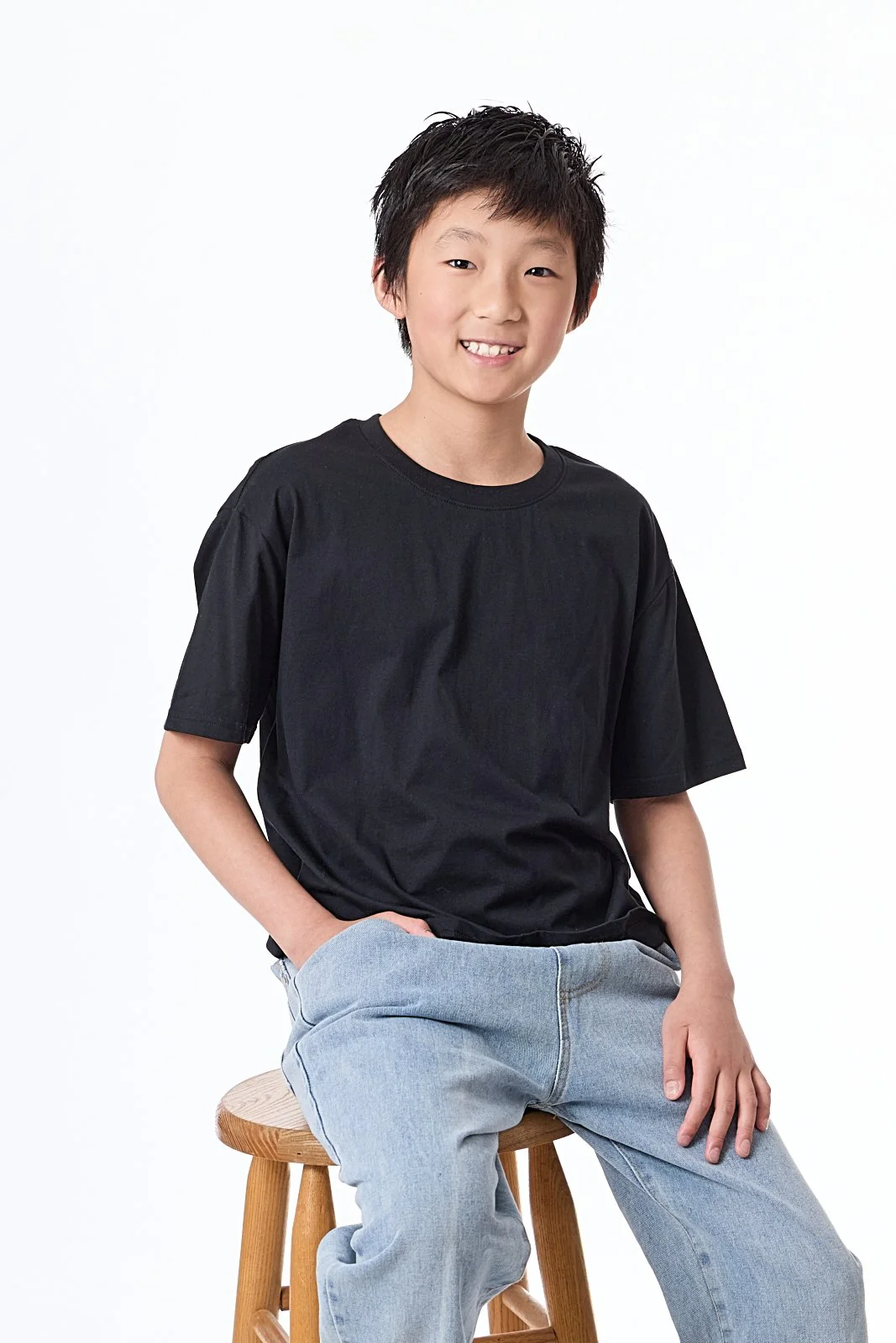 A young boy with black hair sitting on a wooden stool, wearing a black T-shirt and light blue jeans, smiling at the camera against a plain white background.
